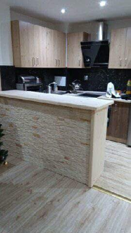 Wykończona kuchnia z drewnianymi szafkami, czarnym blatem i okapem, oraz barkiem wyłożonym dekoracyjną płytką imitującą kamień.