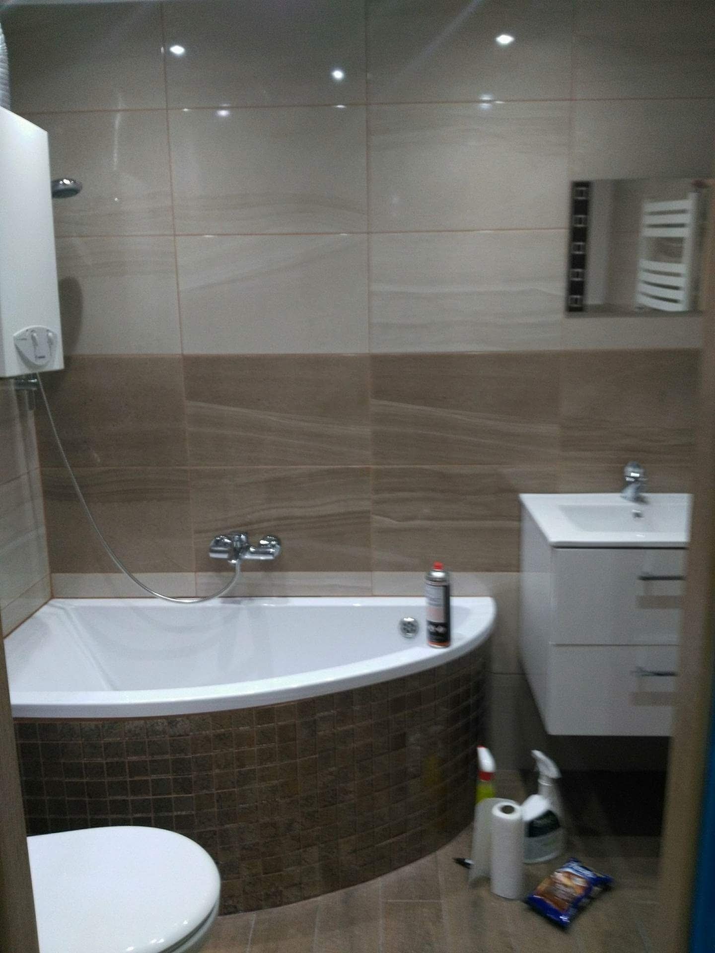 Wykończona łazienka z wanną obudowaną brązową mozaiką, białą umywalką z szafką i grzejnikiem drabinkowym w tle. Na wannie stoi puszka z preparatem, a na podłodze leżą środki czystości i paczka...