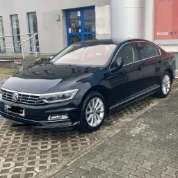 Czarny Volkswagen Passat zaparkowany przed budynkiem z reklamą Bravoauto, widoczny z boku na kostce brukowej.