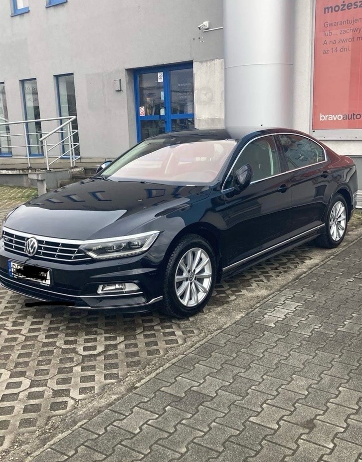 Czarny Volkswagen Passat zaparkowany przed budynkiem z reklamą Bravoauto, widoczny z boku na kostce brukowej.