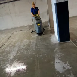 Pracownica w niebieskiej koszulce czyści posadzkę w garażu podziemnym za pomocą szorowarki przemysłowej, widoczne mokre ślady po czyszczeniu, betonowe ściany i słupy.