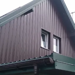 Fragment brązowej elewacji budynku z blachy trapezowej, widoczne okna z białymi ramami, zielone obróbki blacharskie i orynnowanie.