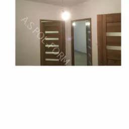 Wnętrze z trzema drewnianymi drzwiami z poziomymi, mlecznymi szybami, widoczna żarówka na kablu, ściany w jasnym odcieniu bieli, logo firmy na ścianie.