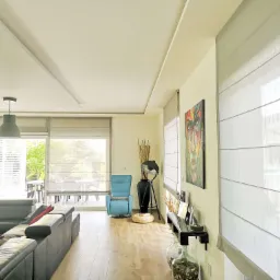 Jasne, przestronne wnętrze salonu z dużymi oknami przesuwnymi, wyposażonymi w kremowe, rzymskie osłony okienne, skórzana sofa i fotel w kolorze turkusowym.