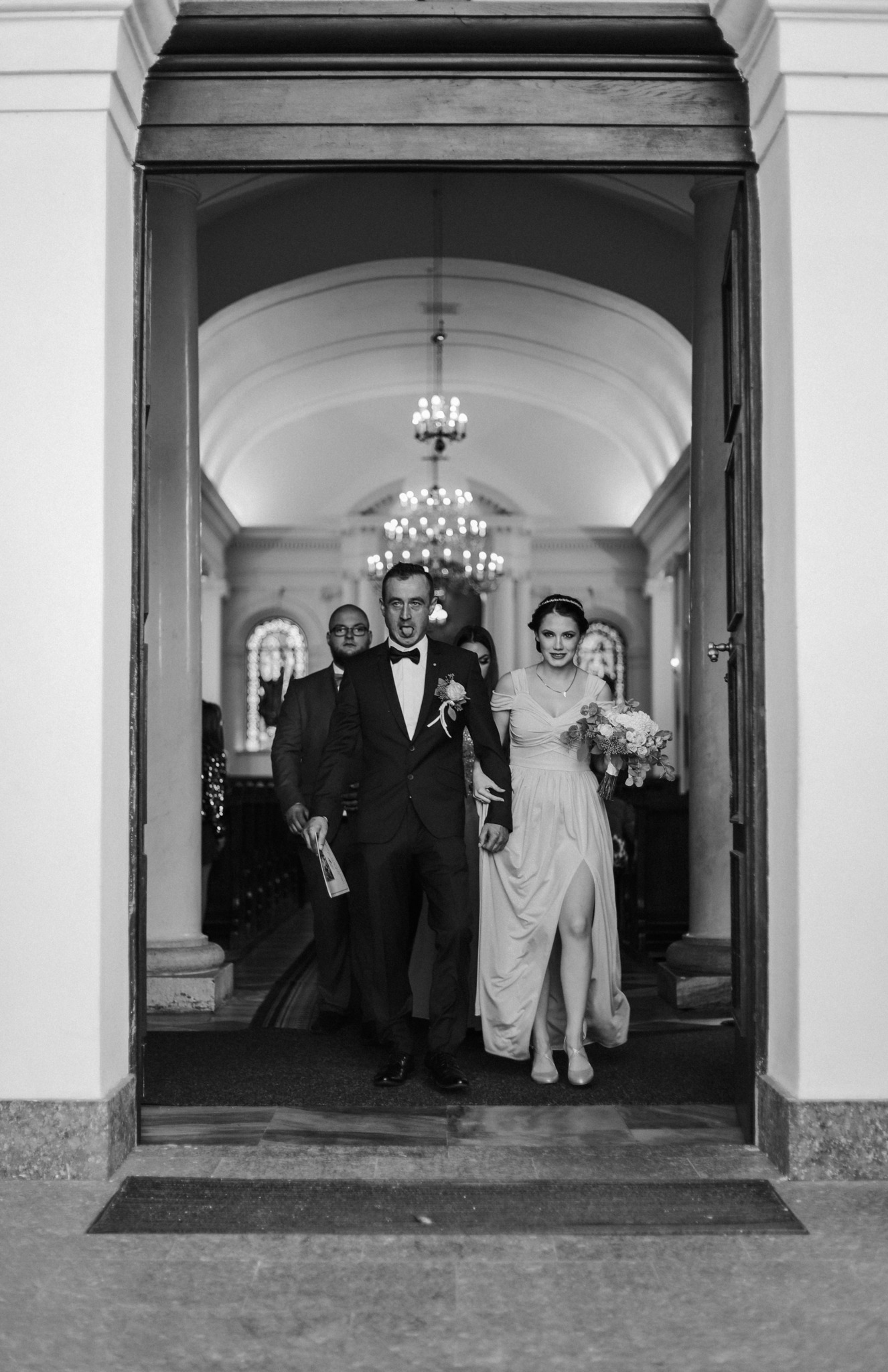 Czarno-białe zdjęcie nowożeńców wychodzących z kościoła przez wysokie, kolumnowe drzwi, za nimi mężczyźni w garniturach, widoczny długi korytarz z żyrandolami.