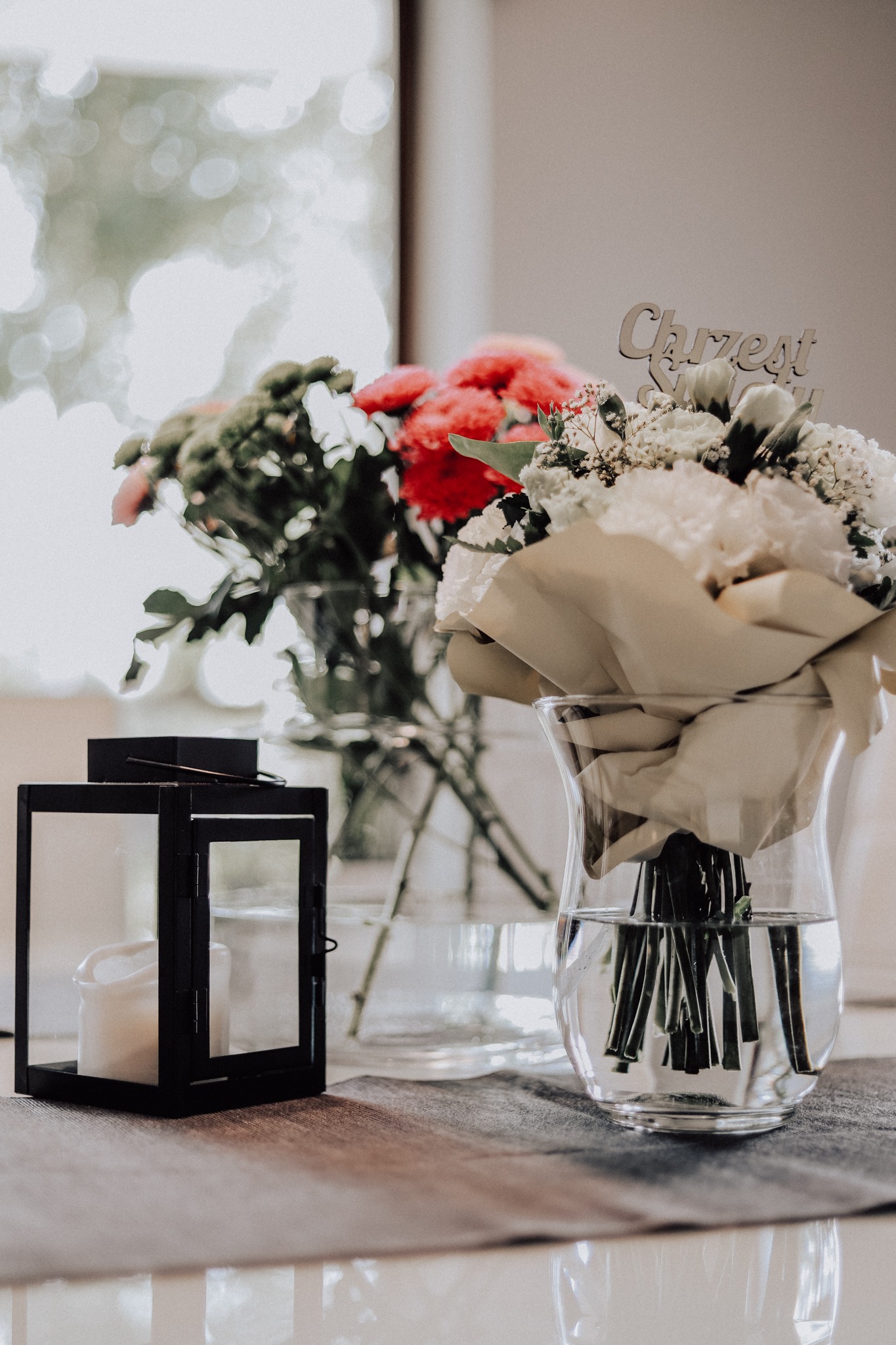 Dekoracja stołu z dwoma bukietami kwiatów w szklanych wazonach oraz czarnym lampionem ze świecą, z napisem 'Chrzest Święty' w bukiecie z przodu.