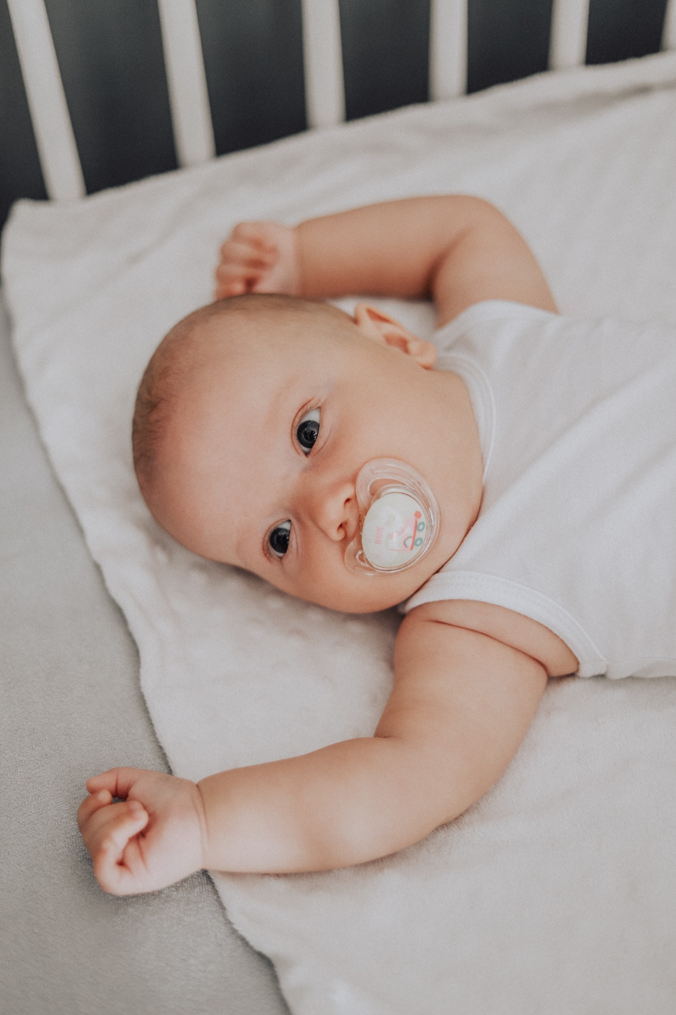 Noworodek leży na plecach w łóżeczku, z otwartymi oczami i smoczkiem w ustach, ubrany w białe body, z rączkami ułożonymi wzdłuż ciała.