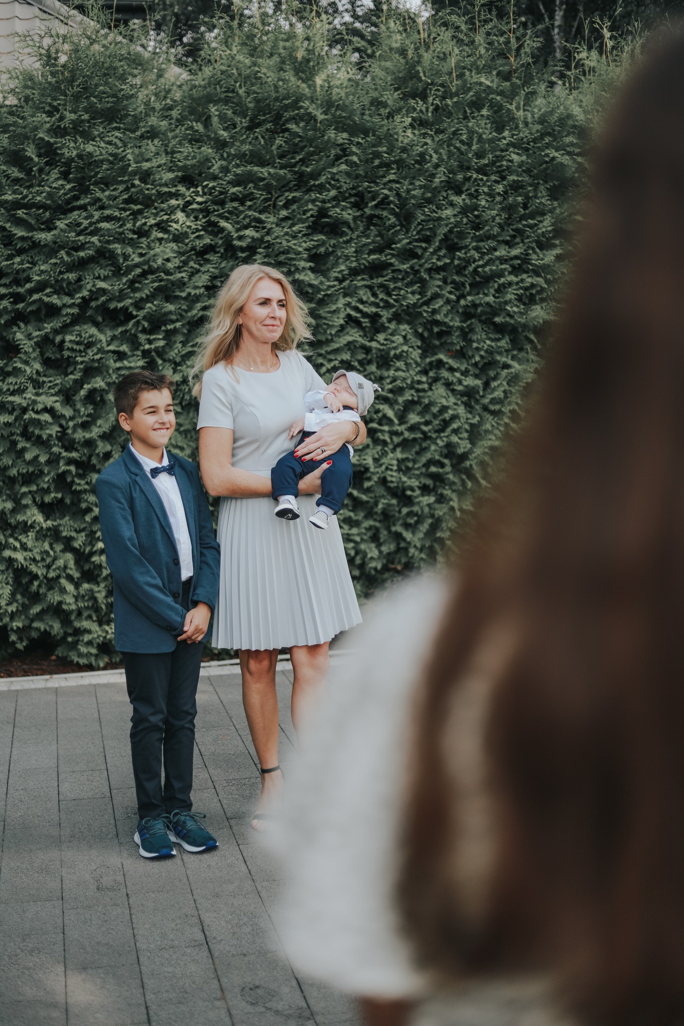 Uśmiechnięta kobieta w szarej sukience trzyma śpiącego niemowlaka w białym ubranku, obok stoi elegancko ubrany chłopiec w niebieskim garniturze, tłem jest zielony żywopłot.