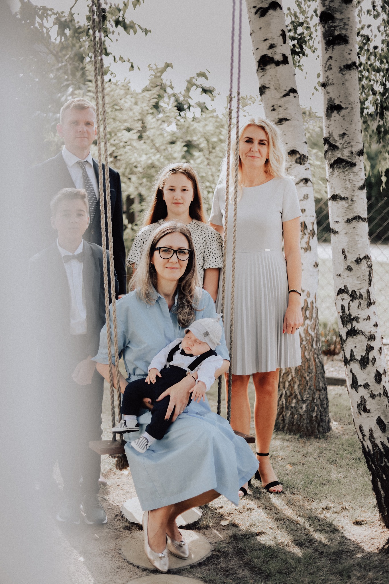Rodzinne zdjęcie plenerowe: mama siedzi na huśtawce z niemowlęciem na kolanach, obok stoi reszta rodziny, w tle drzewa brzozowe.