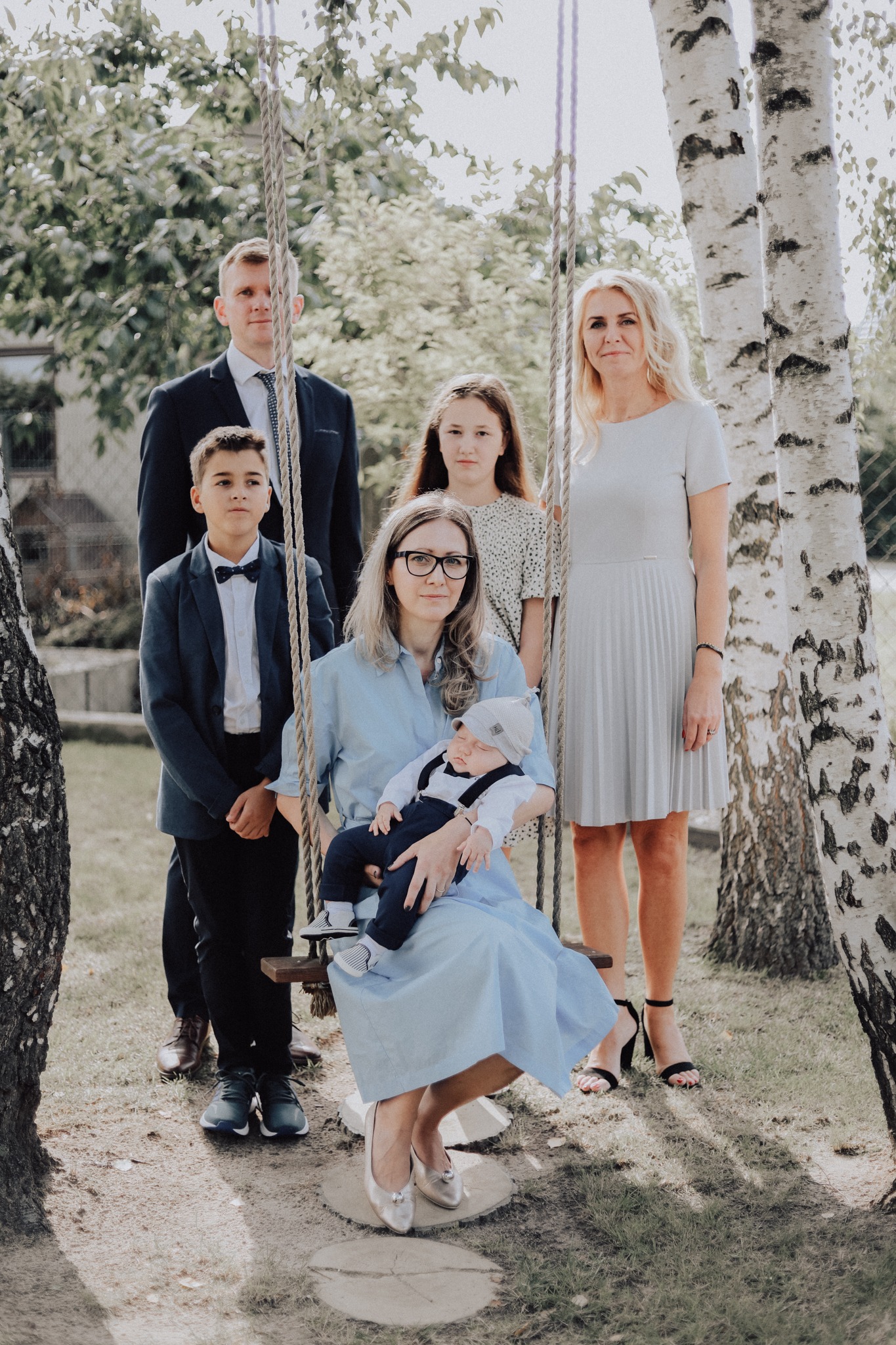 Rodzinne zdjęcie grupowe w plenerze: matka siedząca na huśtawce z niemowlęciem na kolanach, obok ojciec, dwójka dzieci i babcia, wszyscy ubrani w eleganckie stroje, tło stanowią brzozy i zieleń.