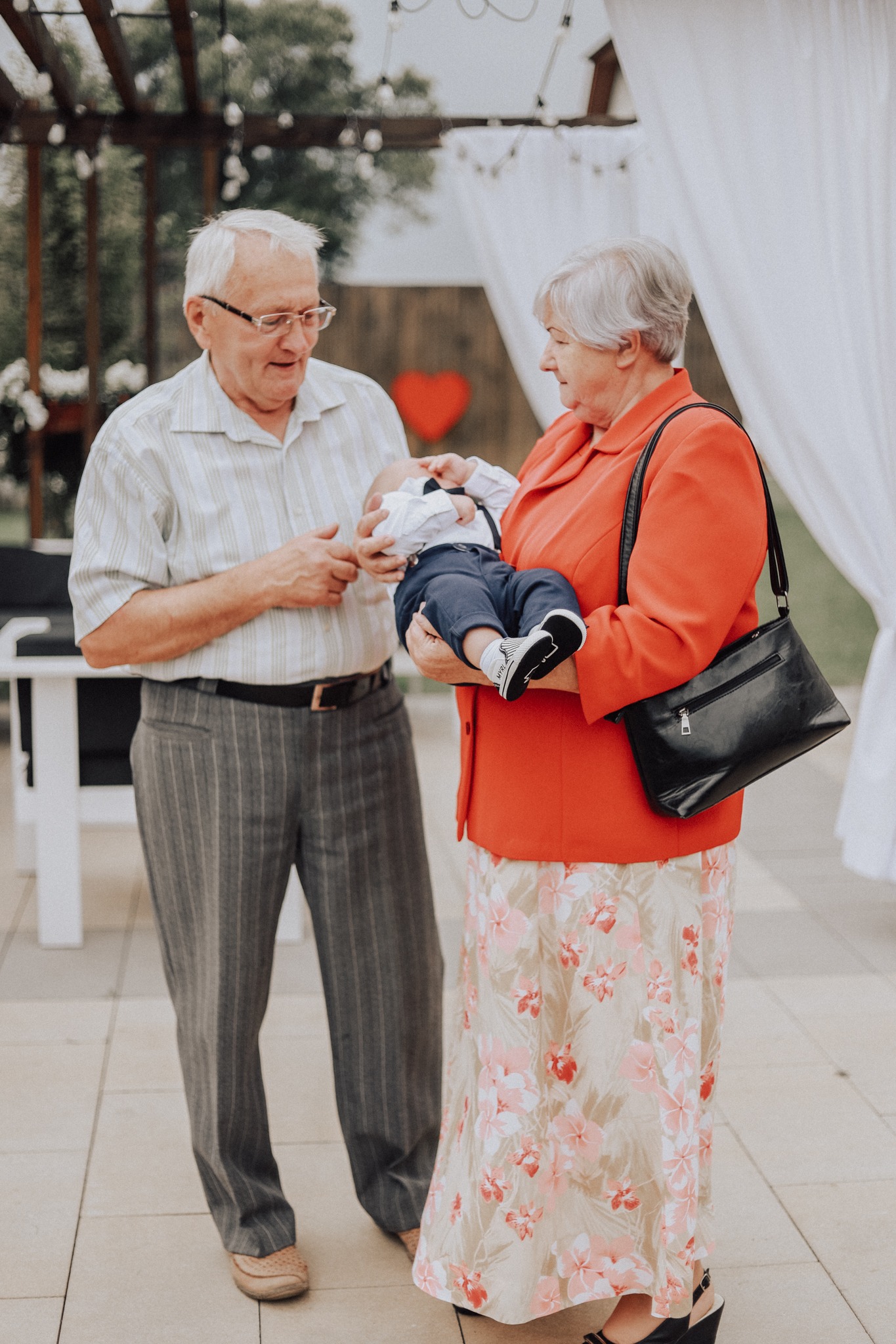 Dziadkowie trzymający noworodka na tle dekoracji z czerwonym sercem i białymi zasłonami.