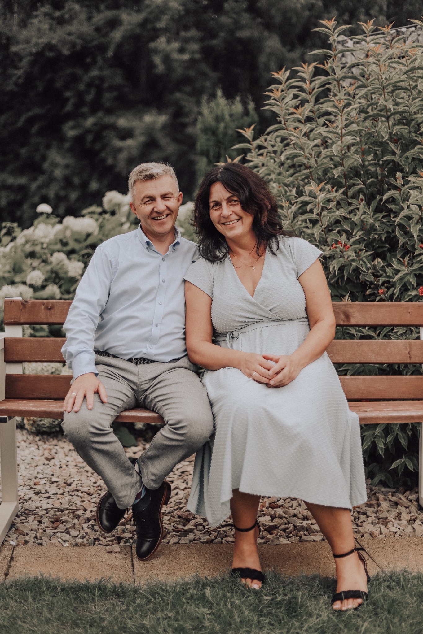 Uśmiechnięta para siedzi na drewnianej ławce w ogrodzie; mężczyzna w błękitnej koszuli i szarych spodniach, kobieta w błękitnej sukience z dekoltem w serek i czarnych sandałach.