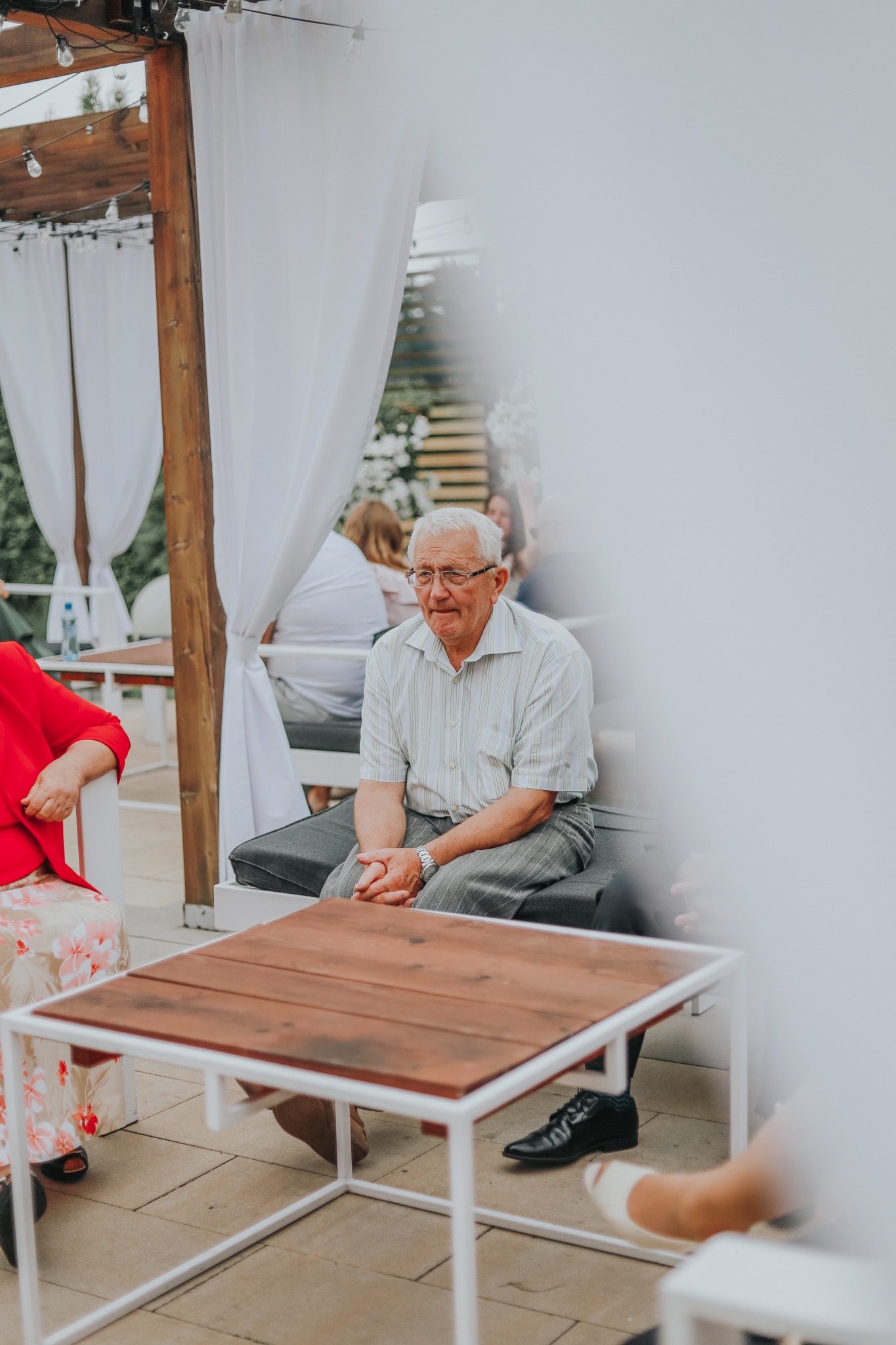 Mężczyzna w okularach siedzi na zewnątrz w Lubartowie, ubrany w koszulę w paski i szare spodnie, z rękami splecionymi na kolanach, w tle białe zasłony i inni ludzie.