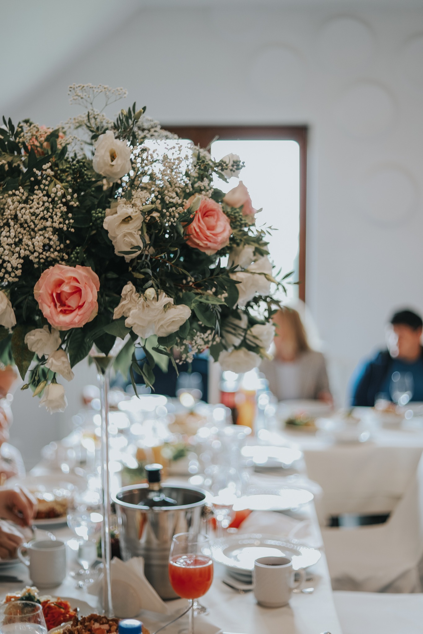 Dekoracja stołu z wysokim bukietem różowych i białych róż oraz gipsówki, srebrny kubełek na lód, kieliszki z czerwonym napojem i talerze z jedzeniem na białym obrusie, w tle siedzący goście.