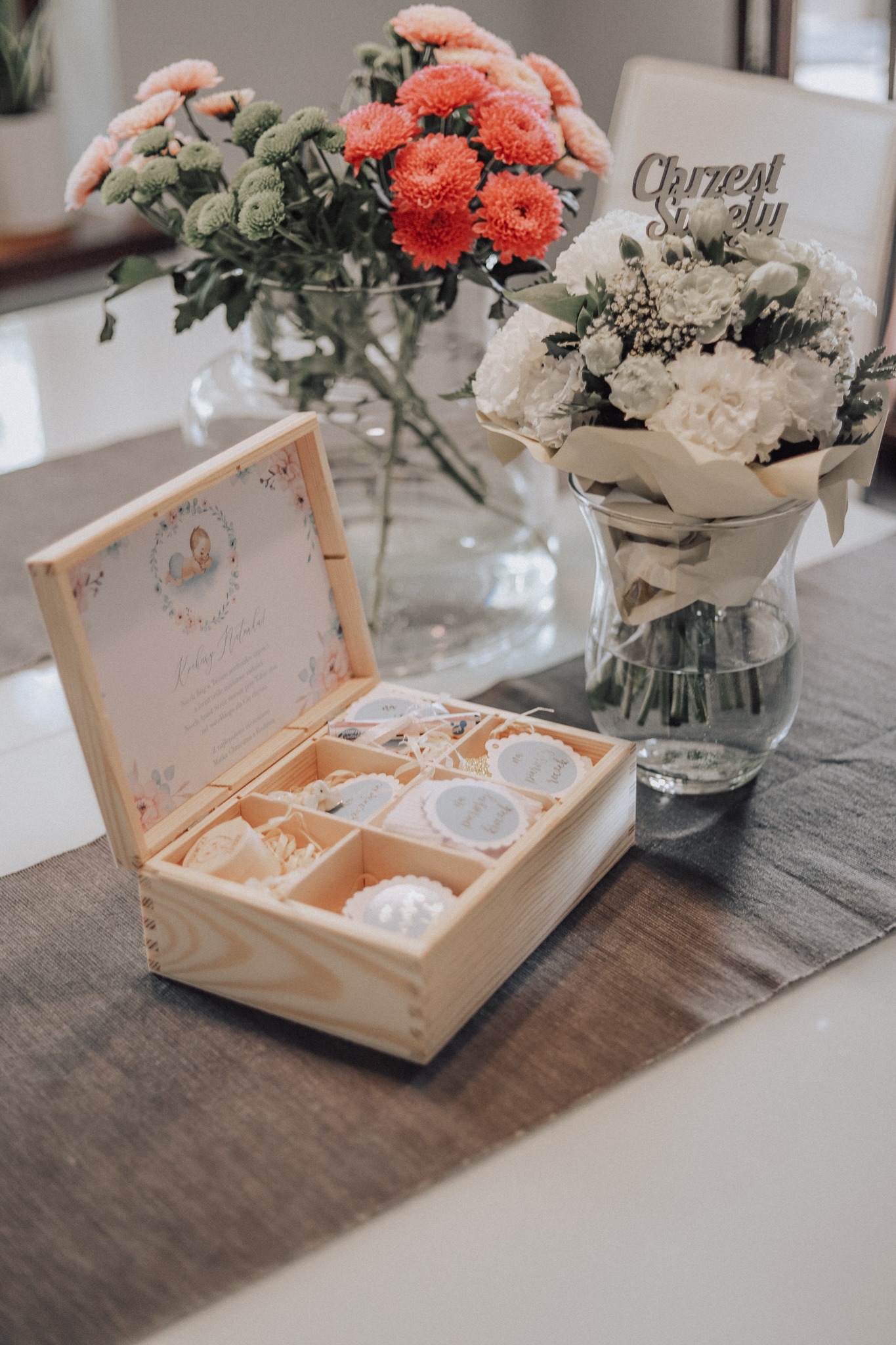 Drewniana skrzynka z przegródkami wypełniona upominkami na chrzest, obok wazony z kwiatami i dekoracja 'Chrzest Święty'.