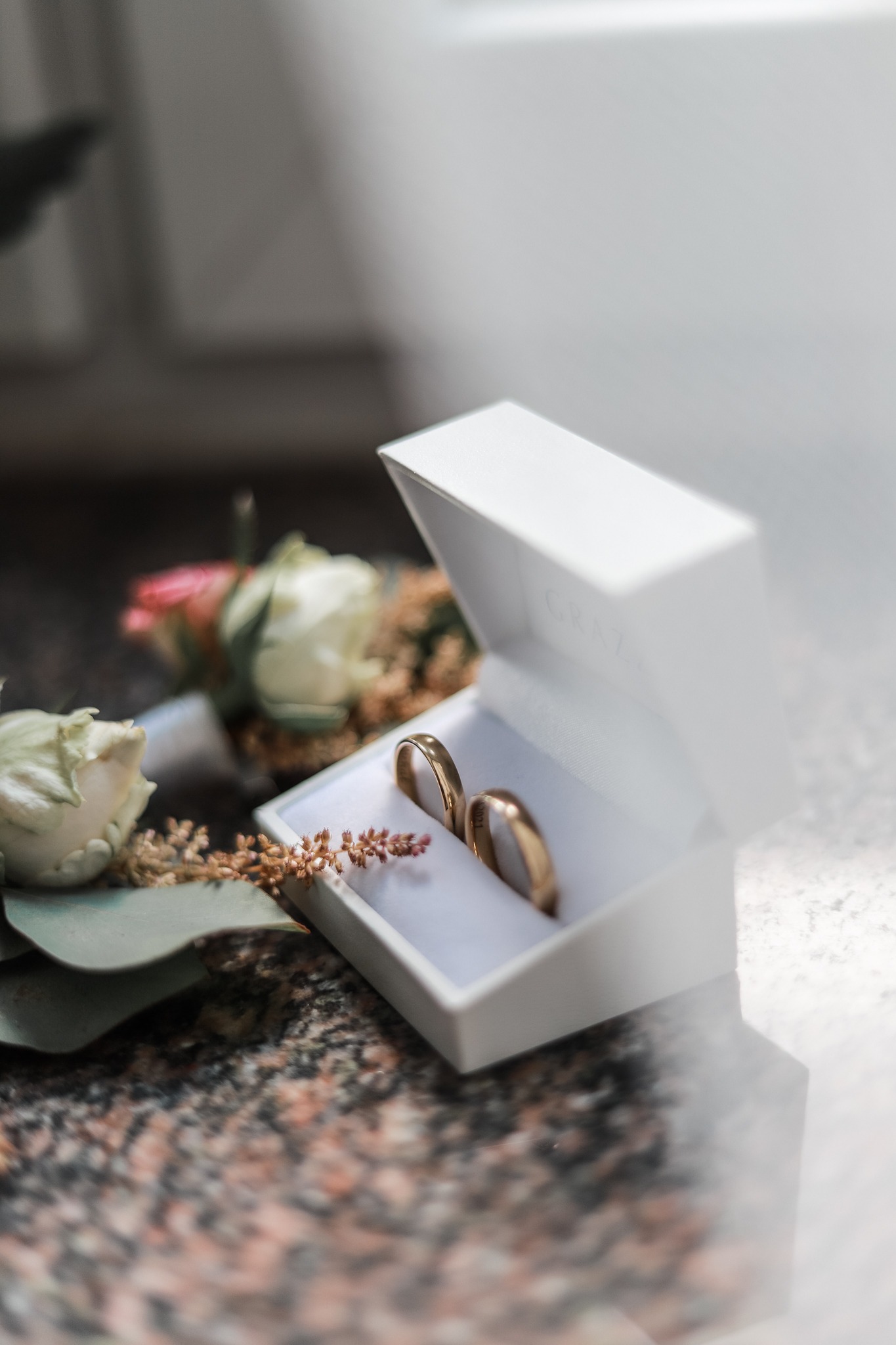 Dwa złote obrączki w białym pudełku z napisem GRAZIA, obok butonierka z białych i różowych róż, na marmurowym blacie.
