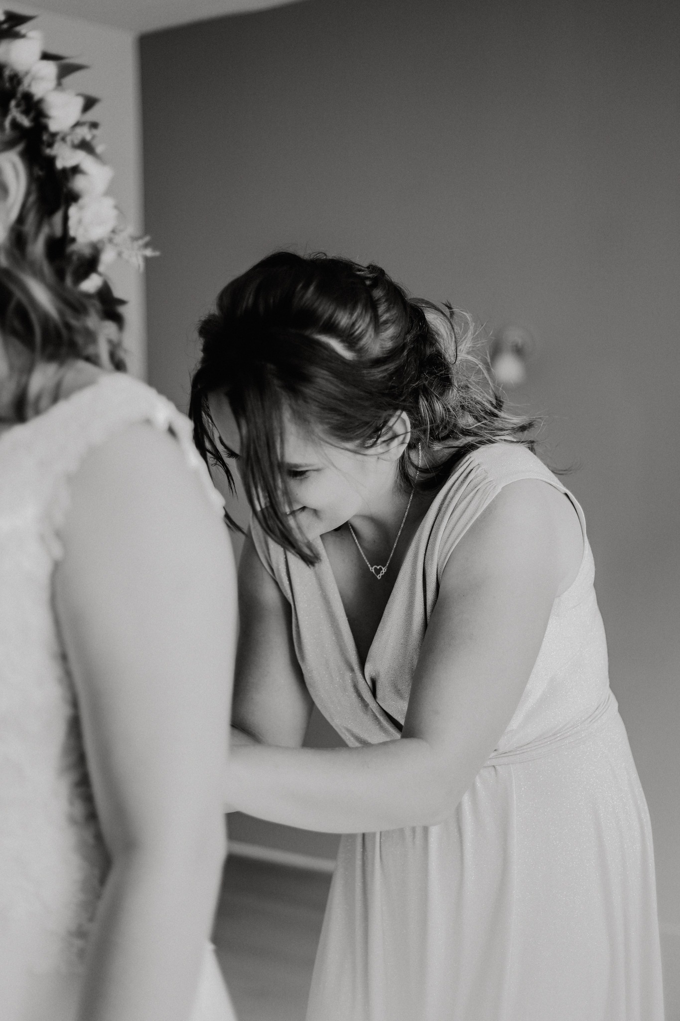 Kobieta w sukience zapina suknię ślubną innej kobiety, widoczny naszyjnik z sercem i kwiatowy wianek na głowie panny młodej, czarno-biała fotografia.