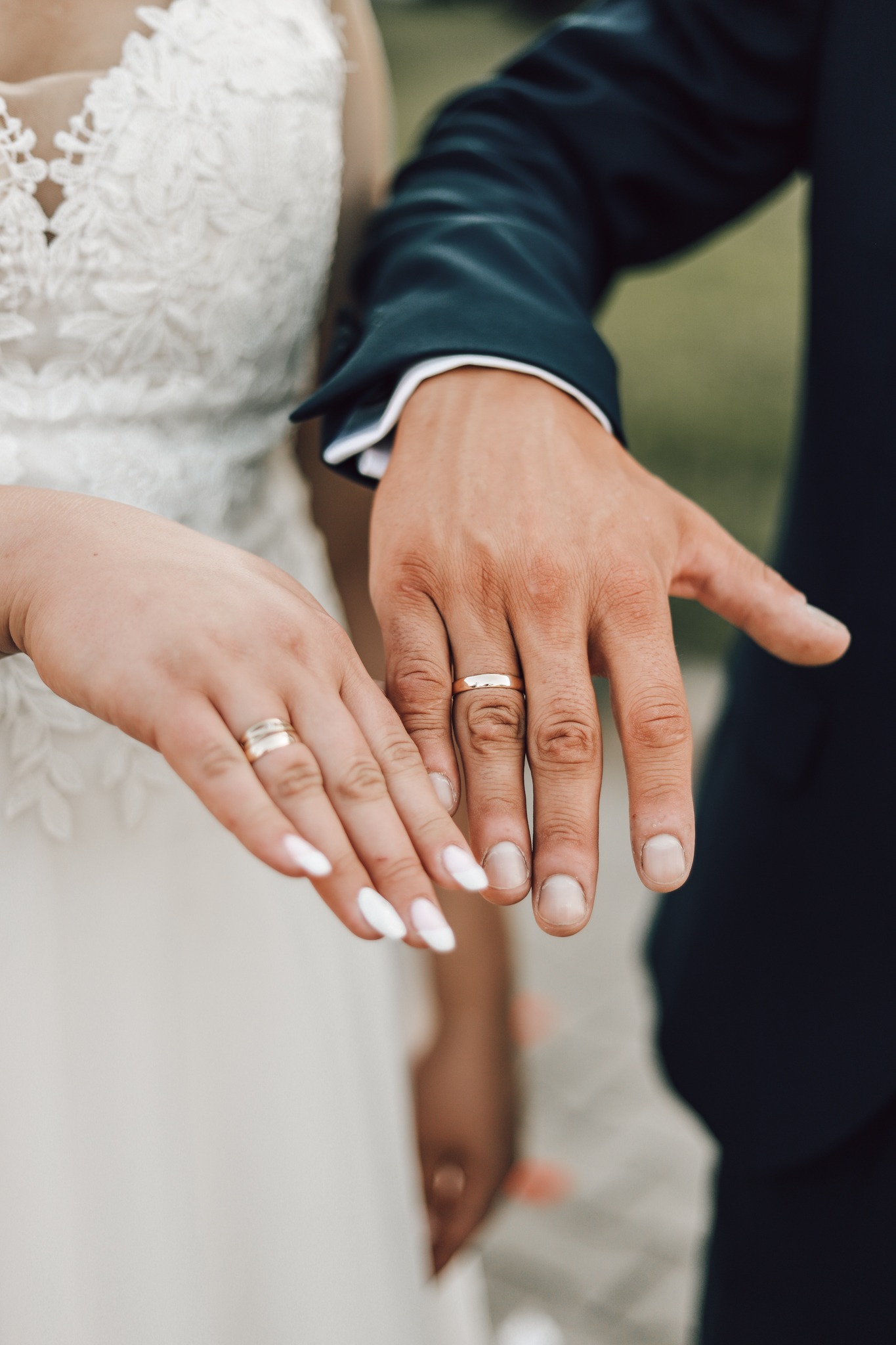 Dłonie nowożeńców z obrączkami ślubnymi; ona w białej sukni z koronką, on w granatowym garniturze.