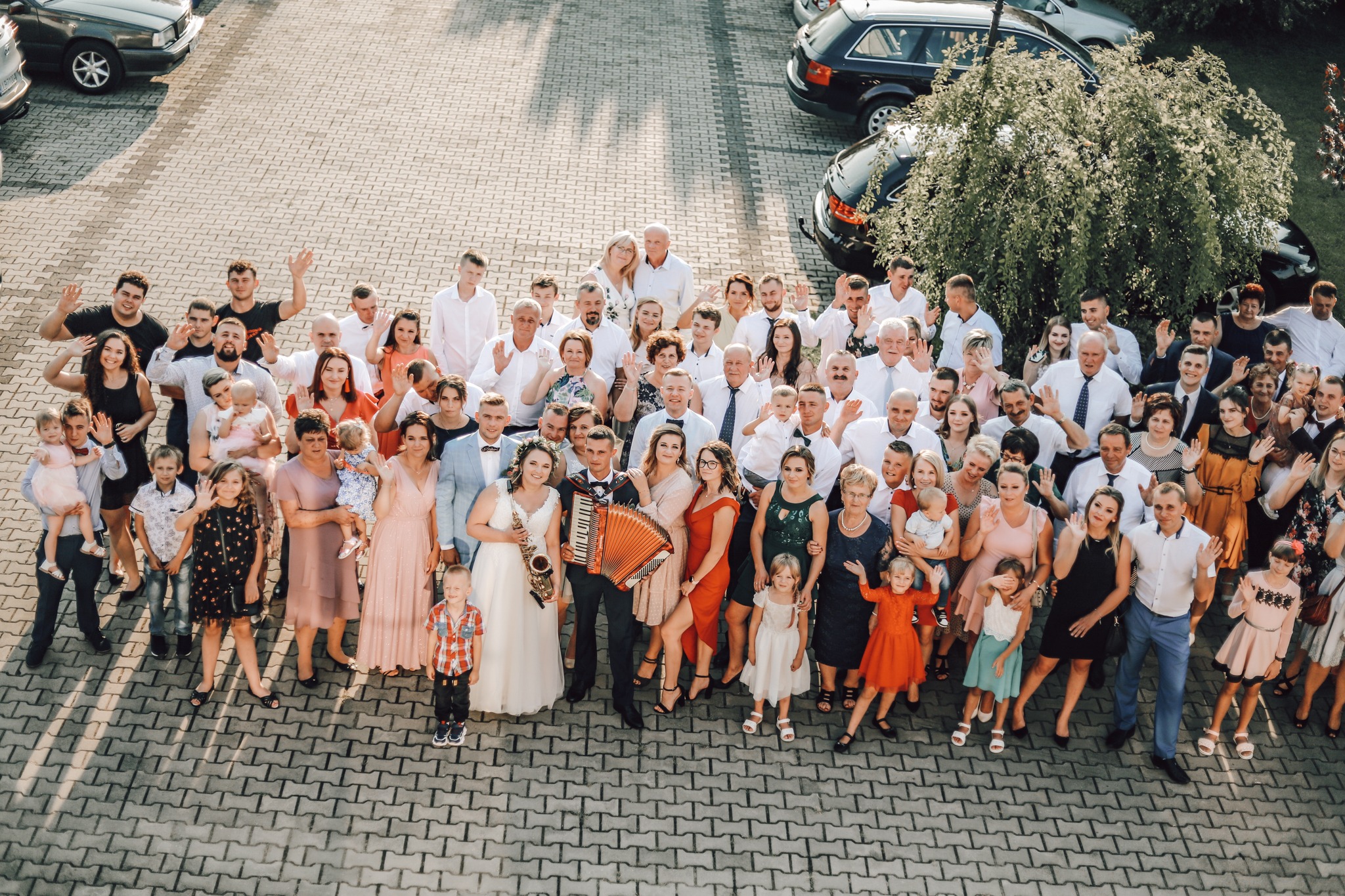 Duża grupa ludzi, w tym para młoda z saksofonem i akordeonem, pozuje do zdjęcia na zewnątrz budynku, wszyscy uśmiechnięci i machający rękami.