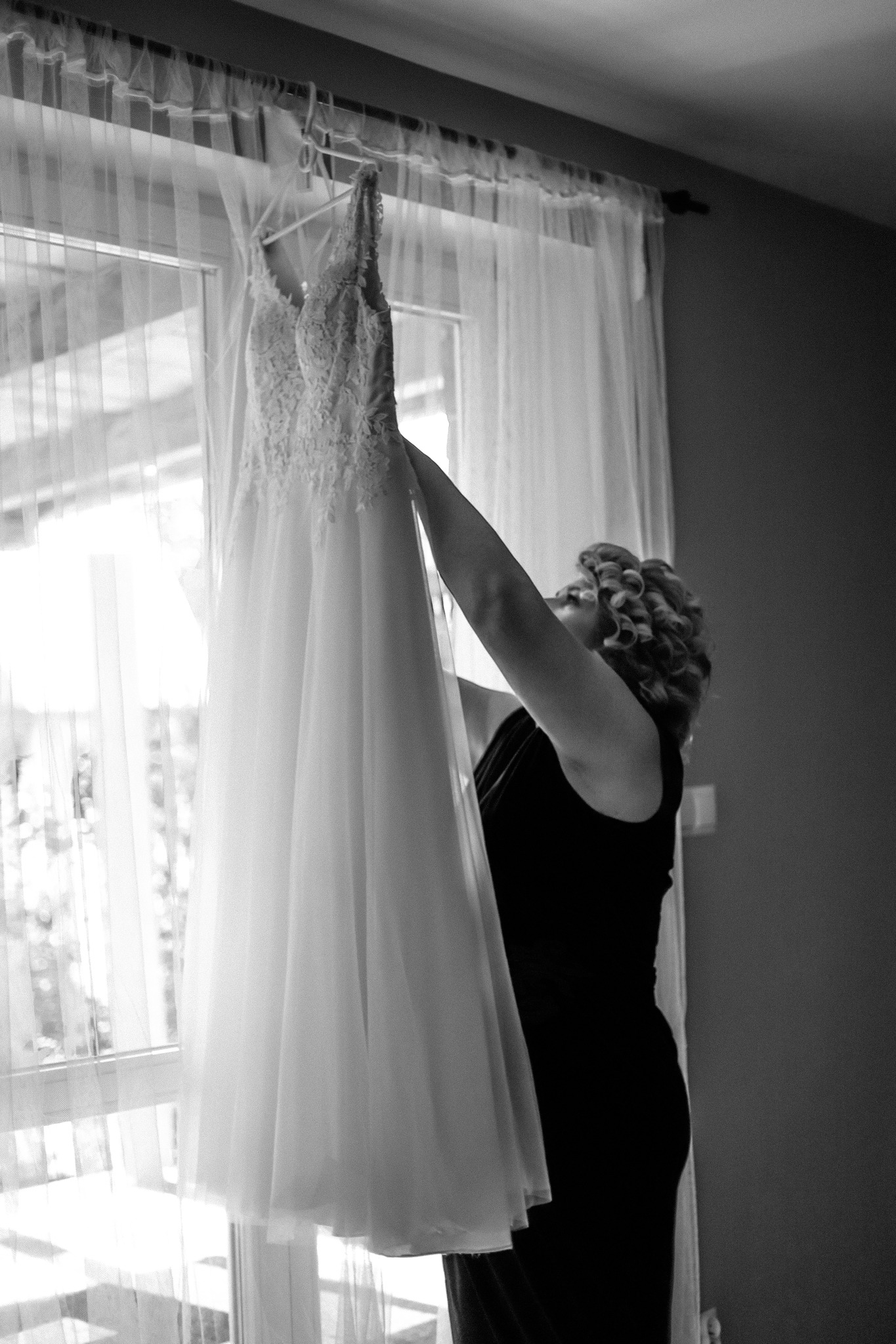 Kobieta w czarnej sukience podnosi białą suknię ślubną z koronkową górą na wieszaku przed oknem z prześwitującymi zasłonami.