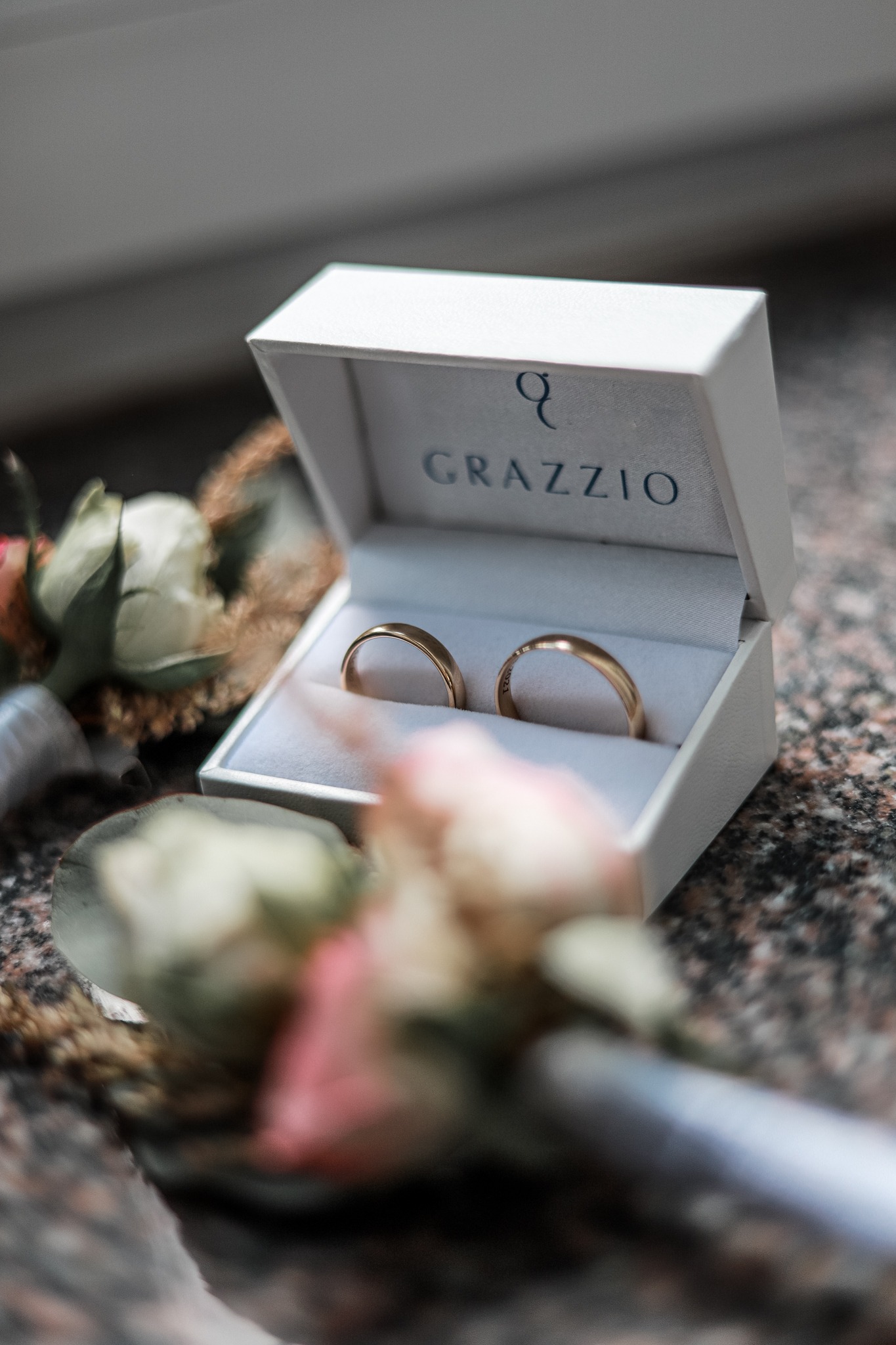 Dwa złote obrączki w białym pudełku firmy Grazzio, obok delikatnych kwiatów w odcieniach bieli i różu, na tle marmurowego blatu.