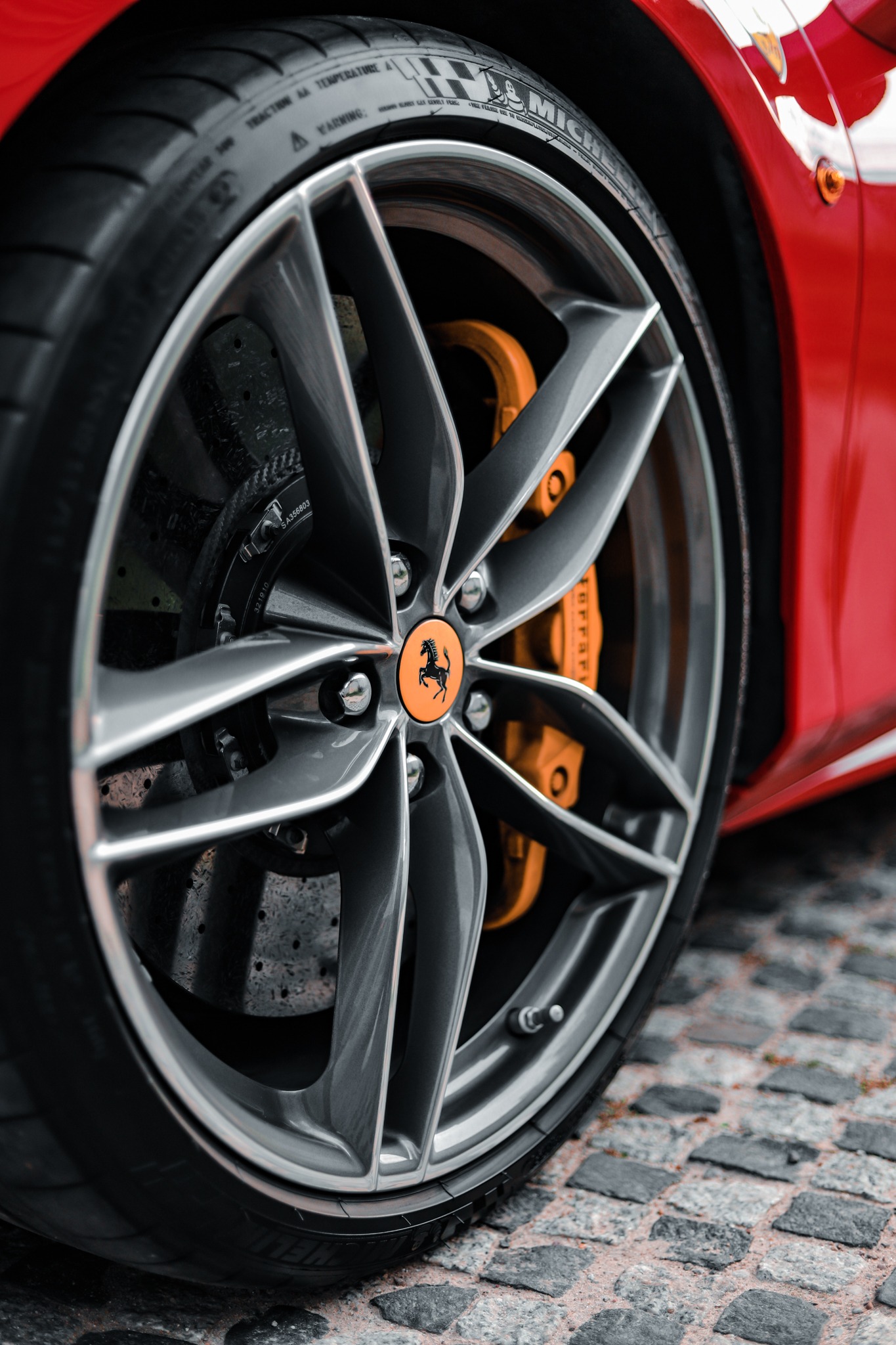 Szczegółowe ujęcie koła sportowego samochodu Ferrari z charakterystycznym logo na pomarańczowym tle, widoczne zaciski hamulcowe i opona na tle kostki brukowej.
