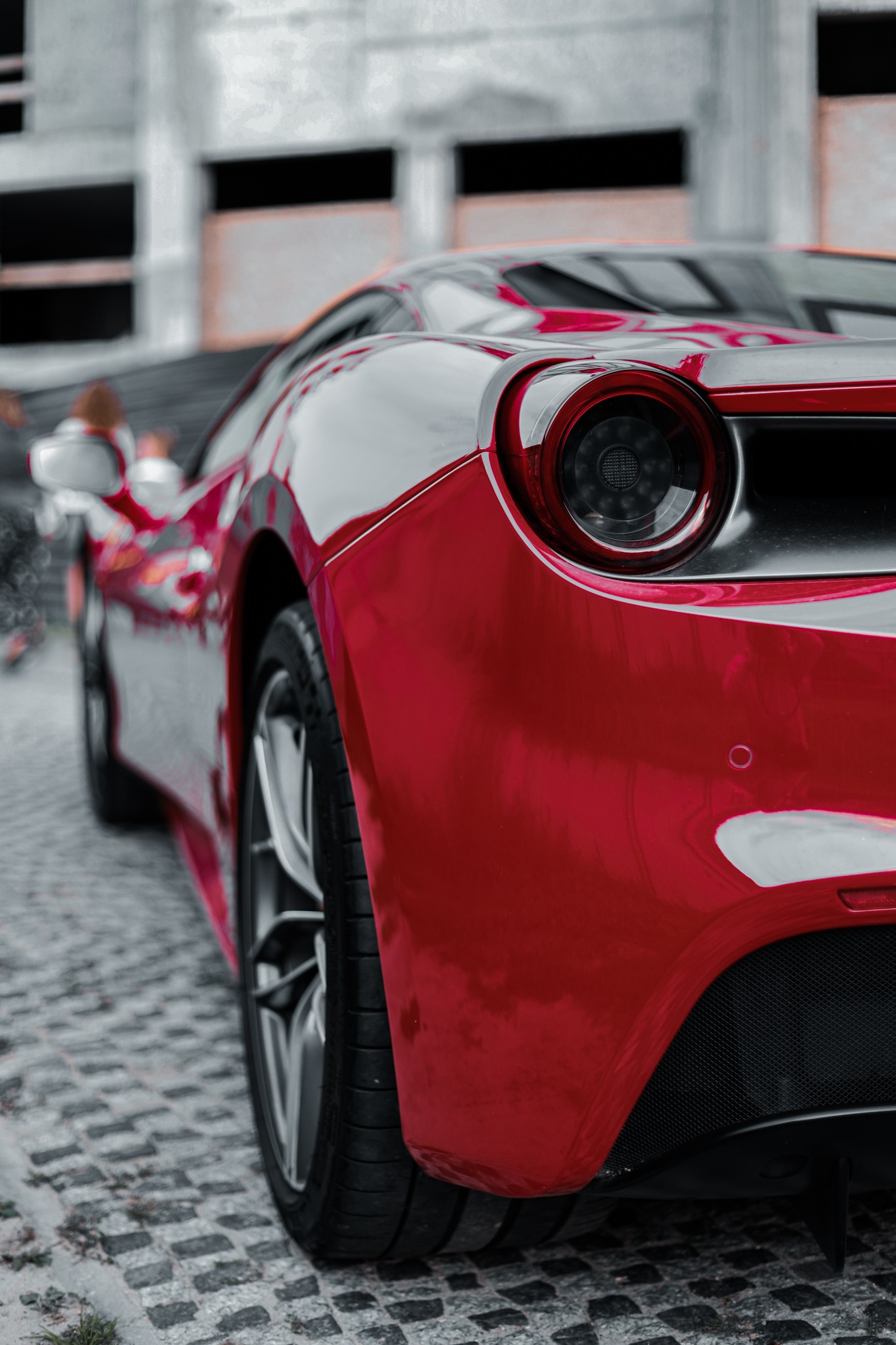 Czerwony sportowy samochód Ferrari, widok z tyłu, na tle szarego, niedokończonego budynku i brukowanej nawierzchni.