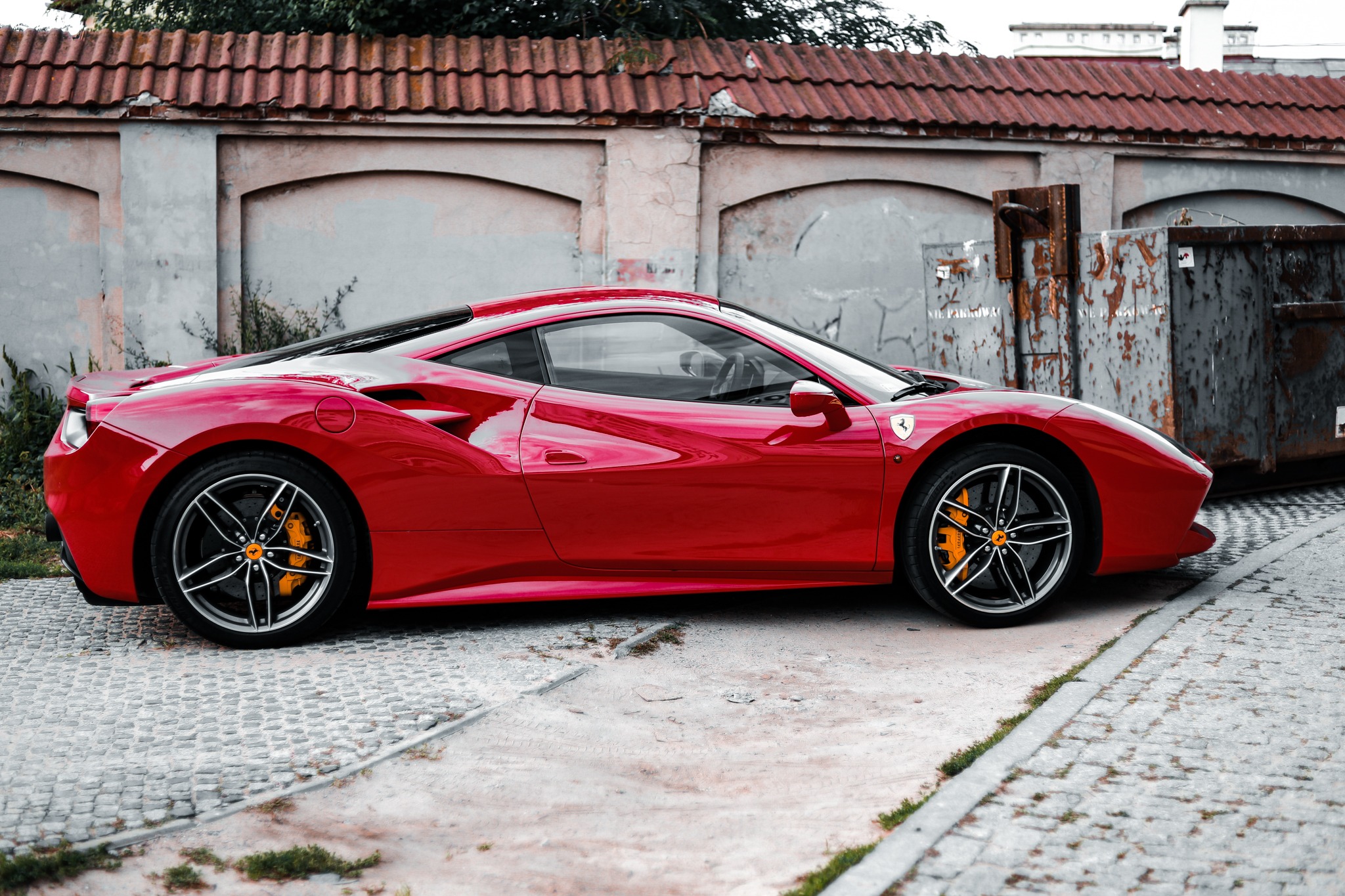 Czerwone Ferrari z żółtymi zaciskami hamulcowymi zaparkowane na brukowanej ulicy, na tle starego muru z dachówką i metalowych kontenerów.