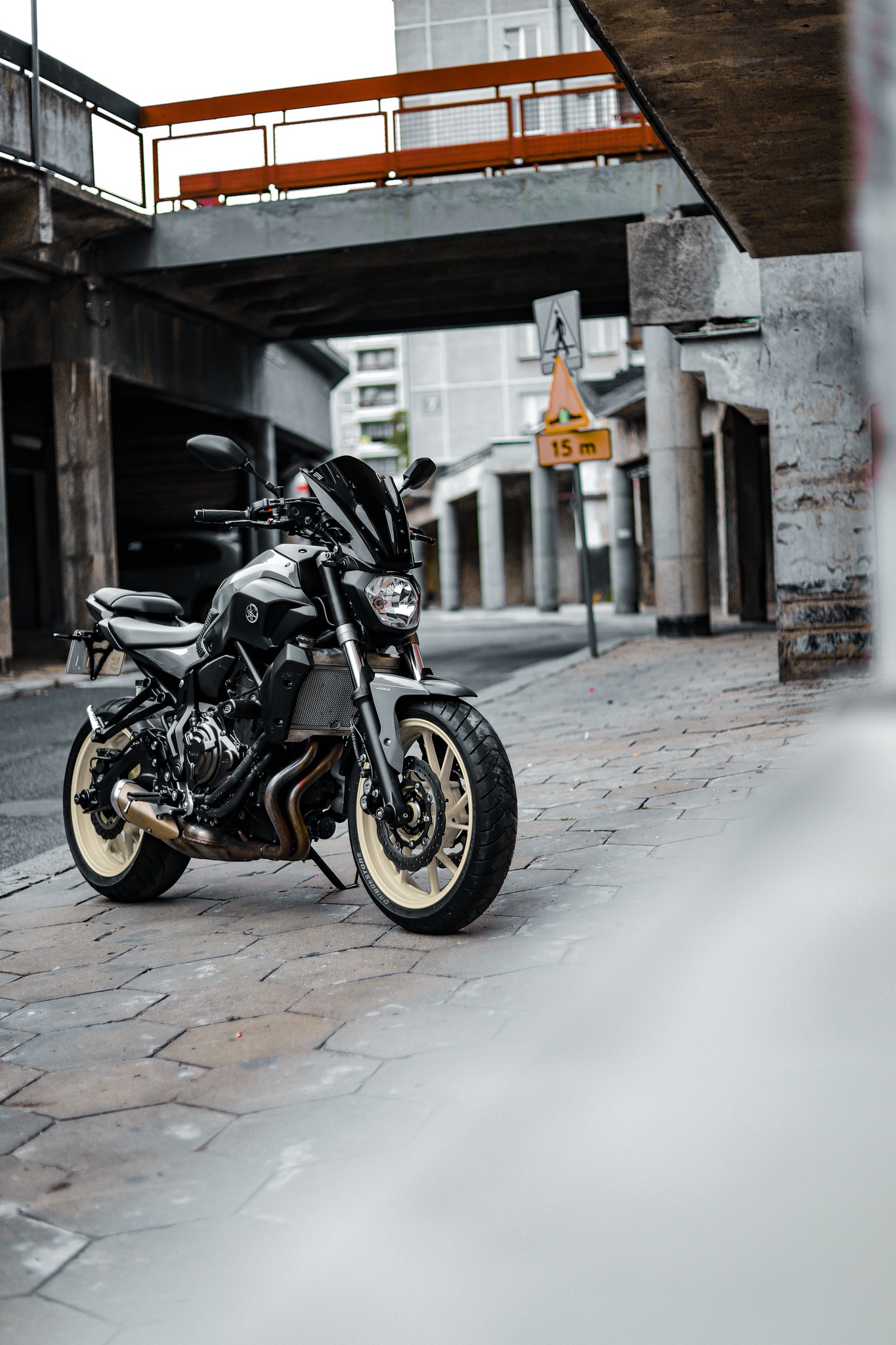 Czarny motocykl Yamaha MT-07 z beżowymi felgami zaparkowany na chodniku z betonowej kostki brukowej, w tle wiadukt i budynki miejskie, znak drogowy informujący o odległości 15 metrów.