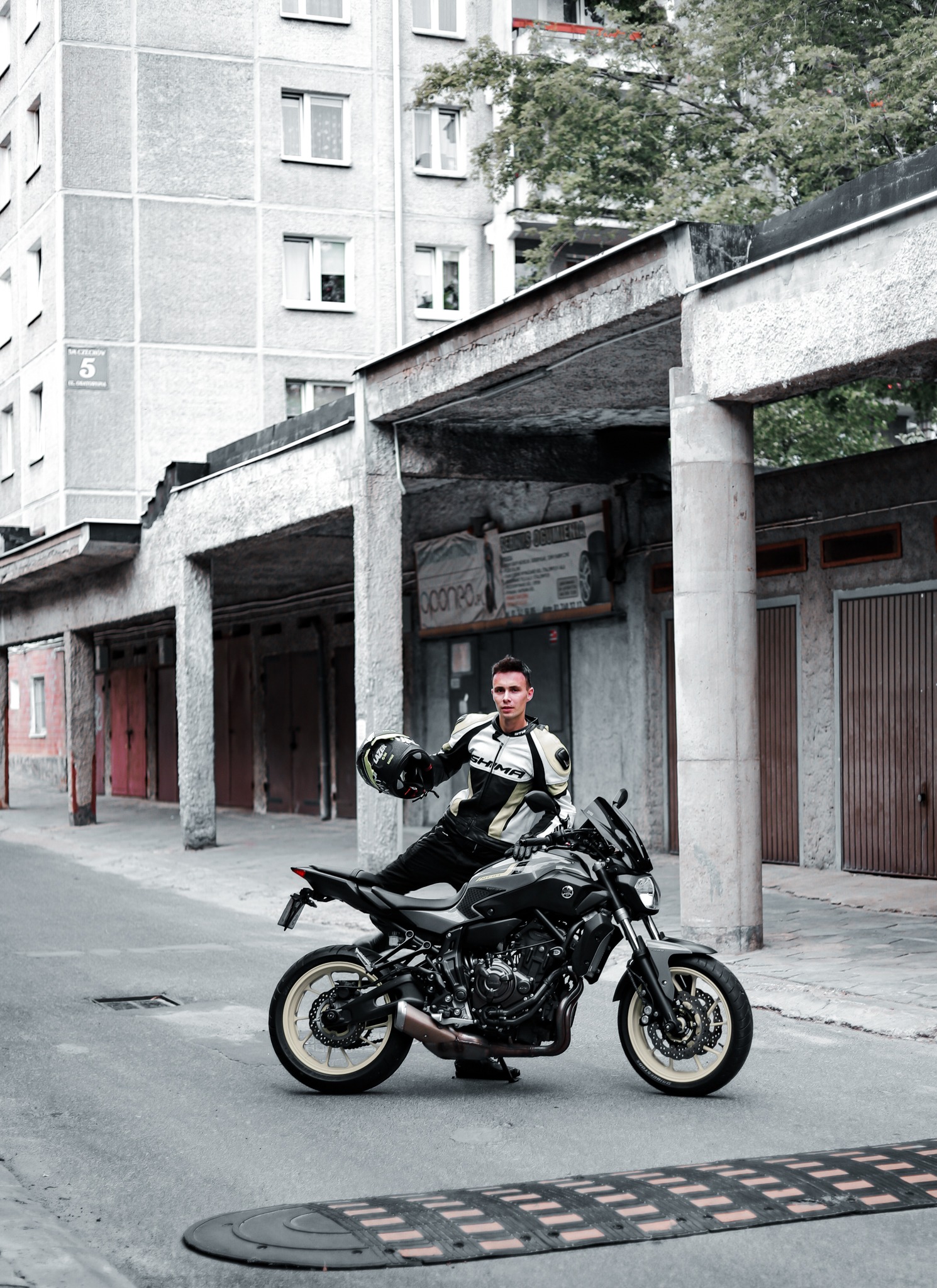 Mężczyzna w kombinezonie motocyklowym siedzi na czarnym motocyklu, trzymając kask na tle szarego bloku mieszkalnego i garaży.