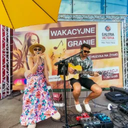 Duet muzyczny na scenie plenerowej: wokalistka w kwiecistej sukience i kapeluszu śpiewa do mikrofonu, gitarzysta gra na gitarze akustycznej, w tle baner reklamowy wydarzenia 'Wakacyjne Granie'...