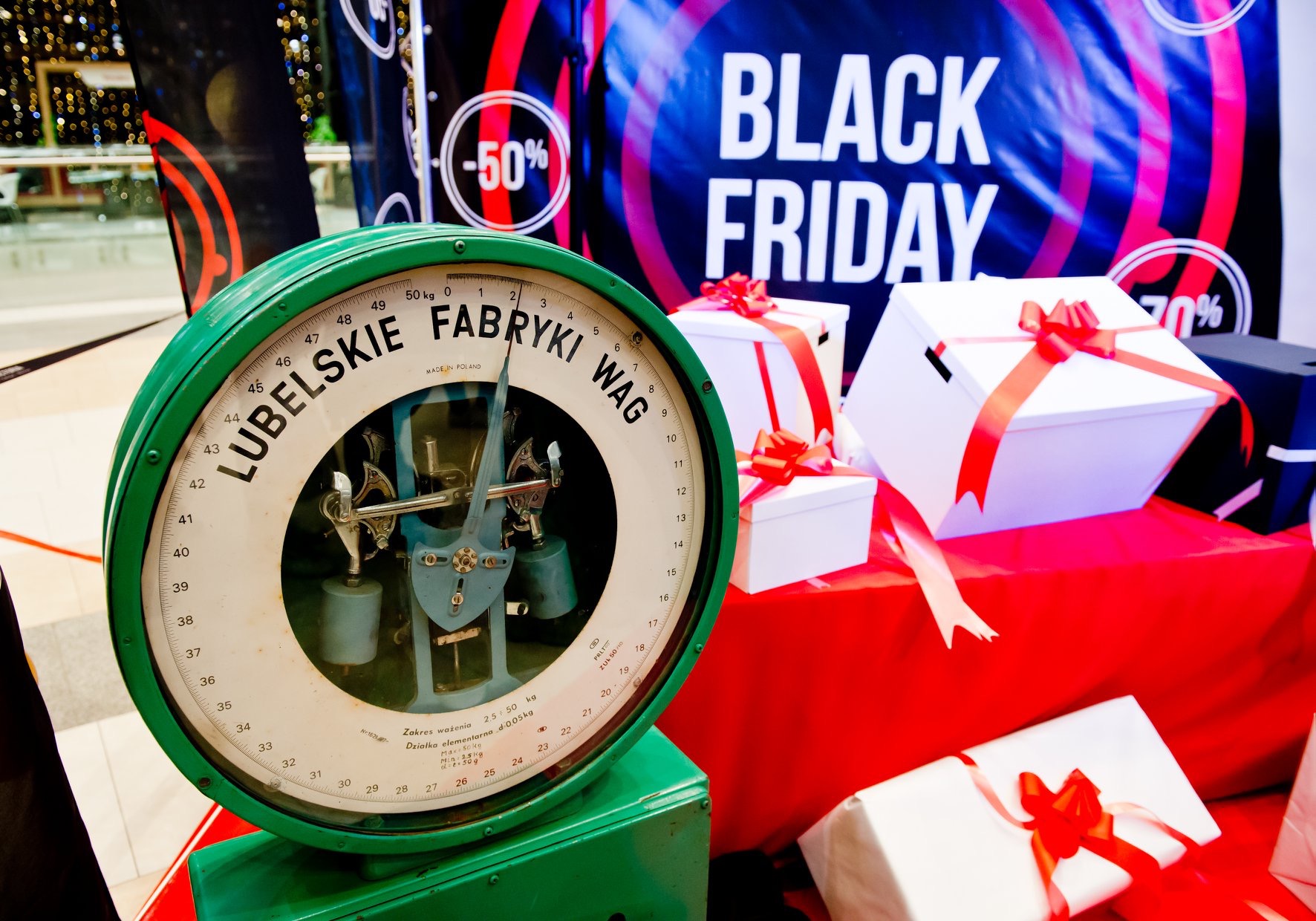 Zielona waga Lubelskie Fabryki Wag na tle białych prezentów z czerwonymi wstążkami i plakatu Black Friday.