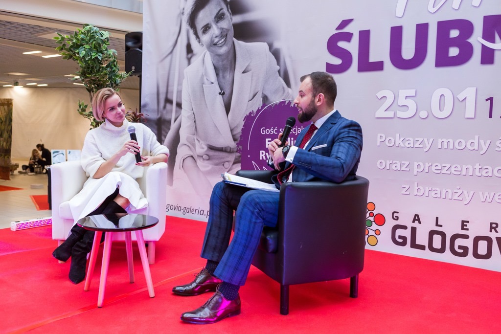 Panel dyskusyjny podczas targów ślubnych; kobieta w białym stroju i mężczyzna w garniturze siedzą w fotelach na czerwonej scenie, rozmawiając do mikrofonów, w tle plakat z napisem 'Ślub'.