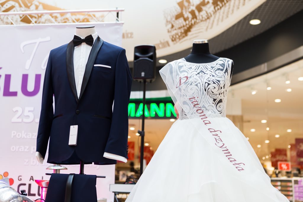 Elegancki granatowy smoking z muchą i biała suknia ślubna z napisem 'Iwona drużmała' na manekinach podczas prezentacji w centrum handlowym, z nagłośnieniem w tle.
