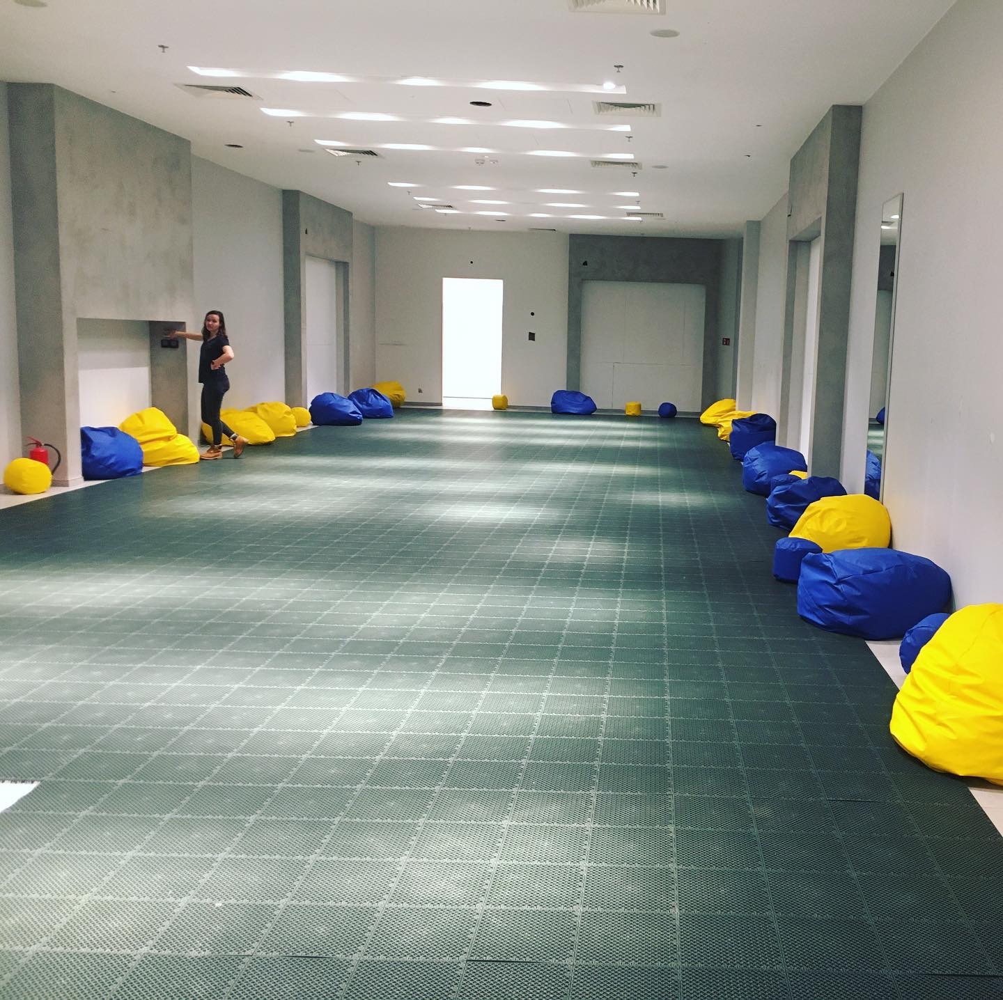 Duża sala z szarymi ścianami i zieloną podłogą wyłożoną kwadratowymi płytami. Wzdłuż ścian ustawione na przemian niebieskie i żółte pufy. Kobieta stoi przy ścianie z gaśnicą obok.