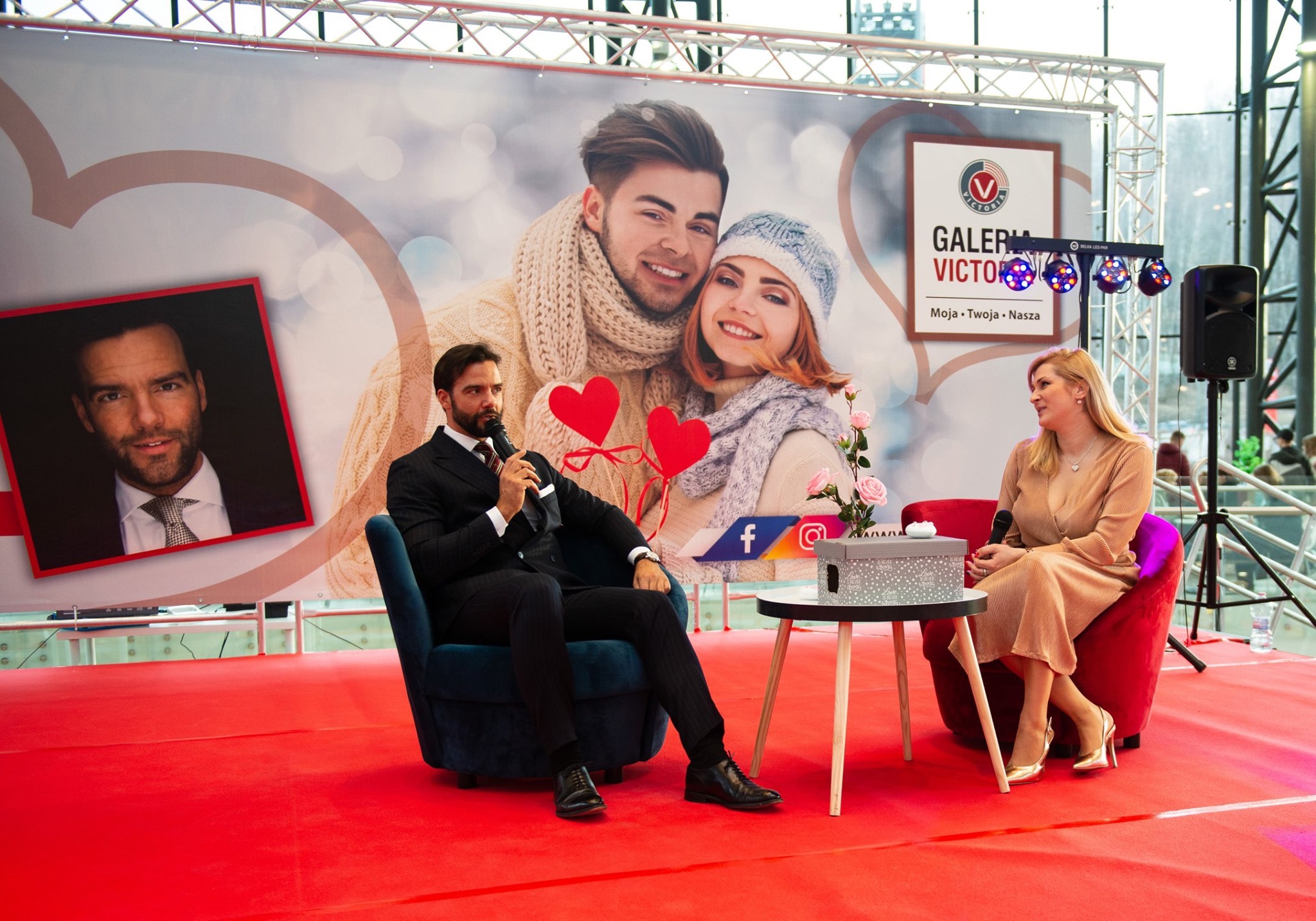 Prezentacja na czerwonej scenie: Mężczyzna w garniturze z mikrofonem i kobieta w sukience siedzą w fotelach na tle baneru reklamowego z parą i logo galerii.