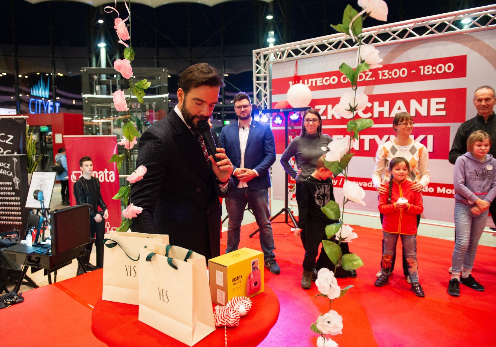 Prezenter w garniturze przemawia do mikrofonu na czerwonej scenie podczas wydarzenia 'Zkochane Entytety' 1 lutego, z torbami prezentowymi YES i aparatem Instax Mini 9 na stole, w tle publiczność...