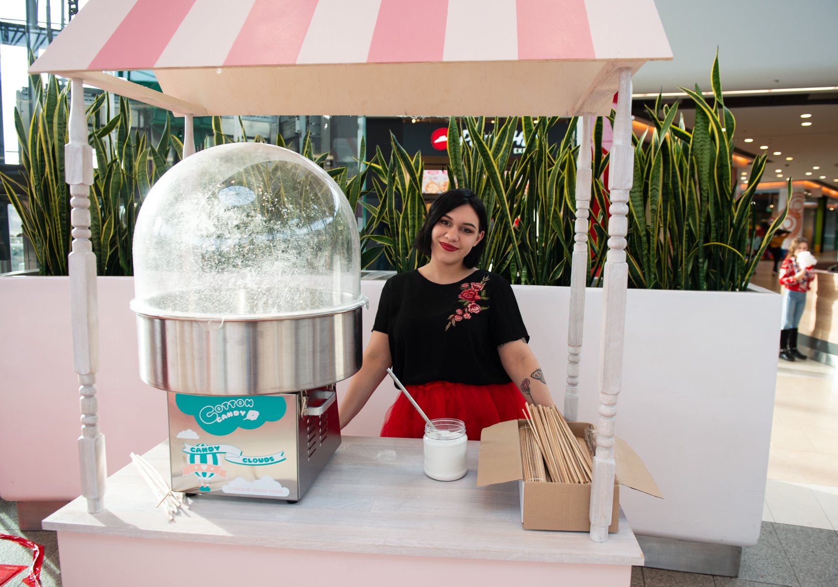 Kobieta za stoiskiem z watą cukrową w centrum handlowym, maszyna do waty cukrowej z napisem 'Cotton Candy Clouds', różowo-biały daszek nad stoiskiem, w tle zielone rośliny w donicach.