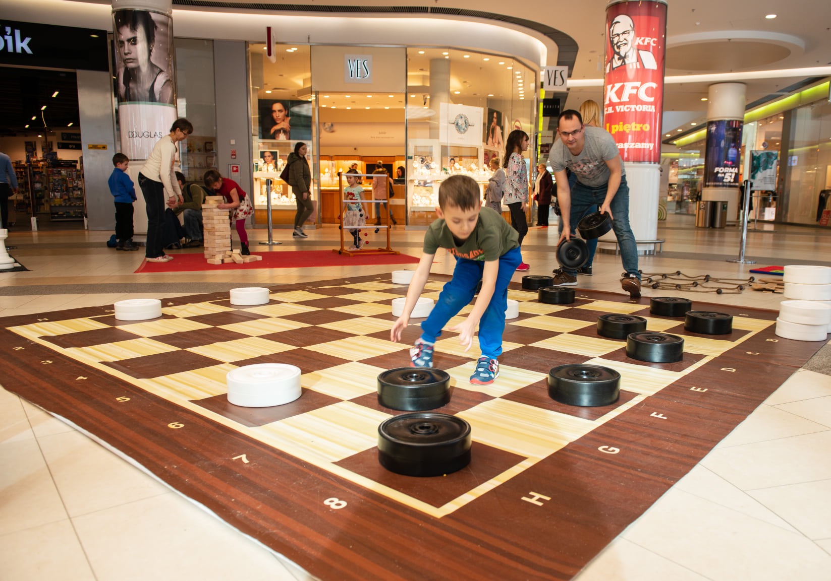 Chłopiec w niebieskich spodniach gra w powiększone warcaby na planszy w centrum handlowym, w tle inni ludzie i sklepy.