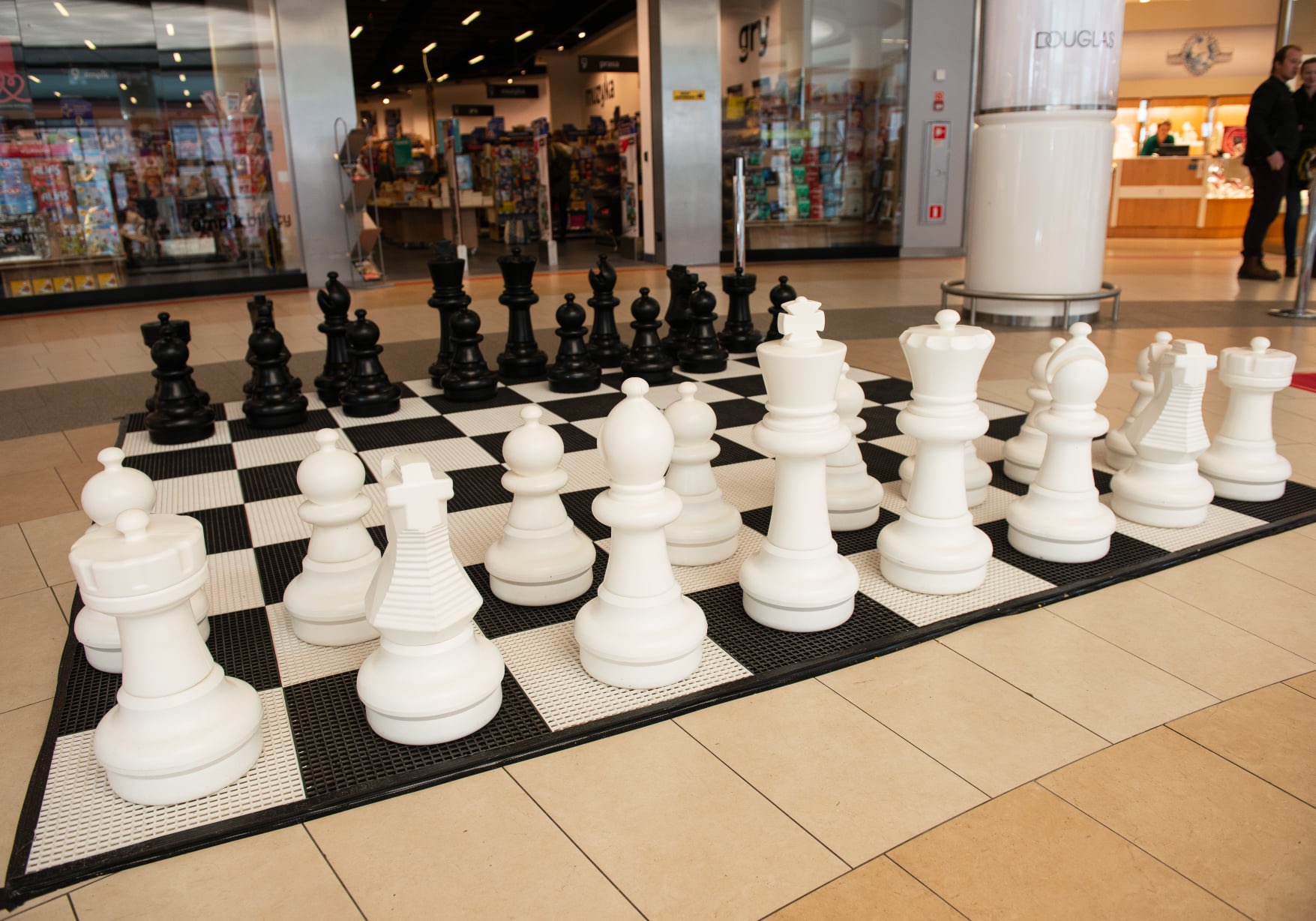 Duża plenerowa szachownica z białymi i czarnymi figurami w centrum handlowym, widoczne sklepy w tle.