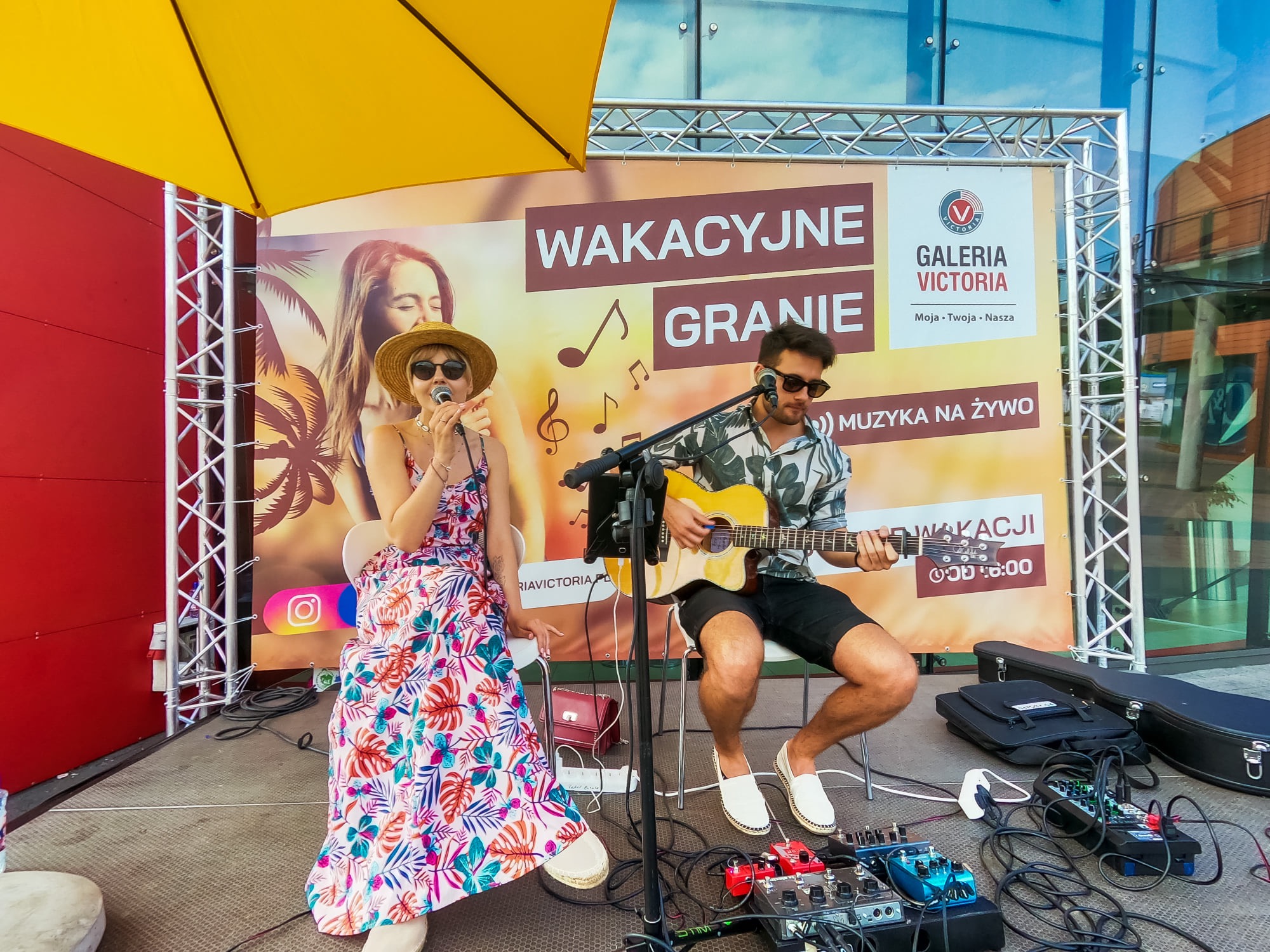 Duet muzyczny na scenie plenerowej: wokalistka w kwiecistej sukience i kapeluszu śpiewa do mikrofonu, gitarzysta gra na gitarze akustycznej, w tle baner reklamowy wydarzenia 'Wakacyjne Granie'...