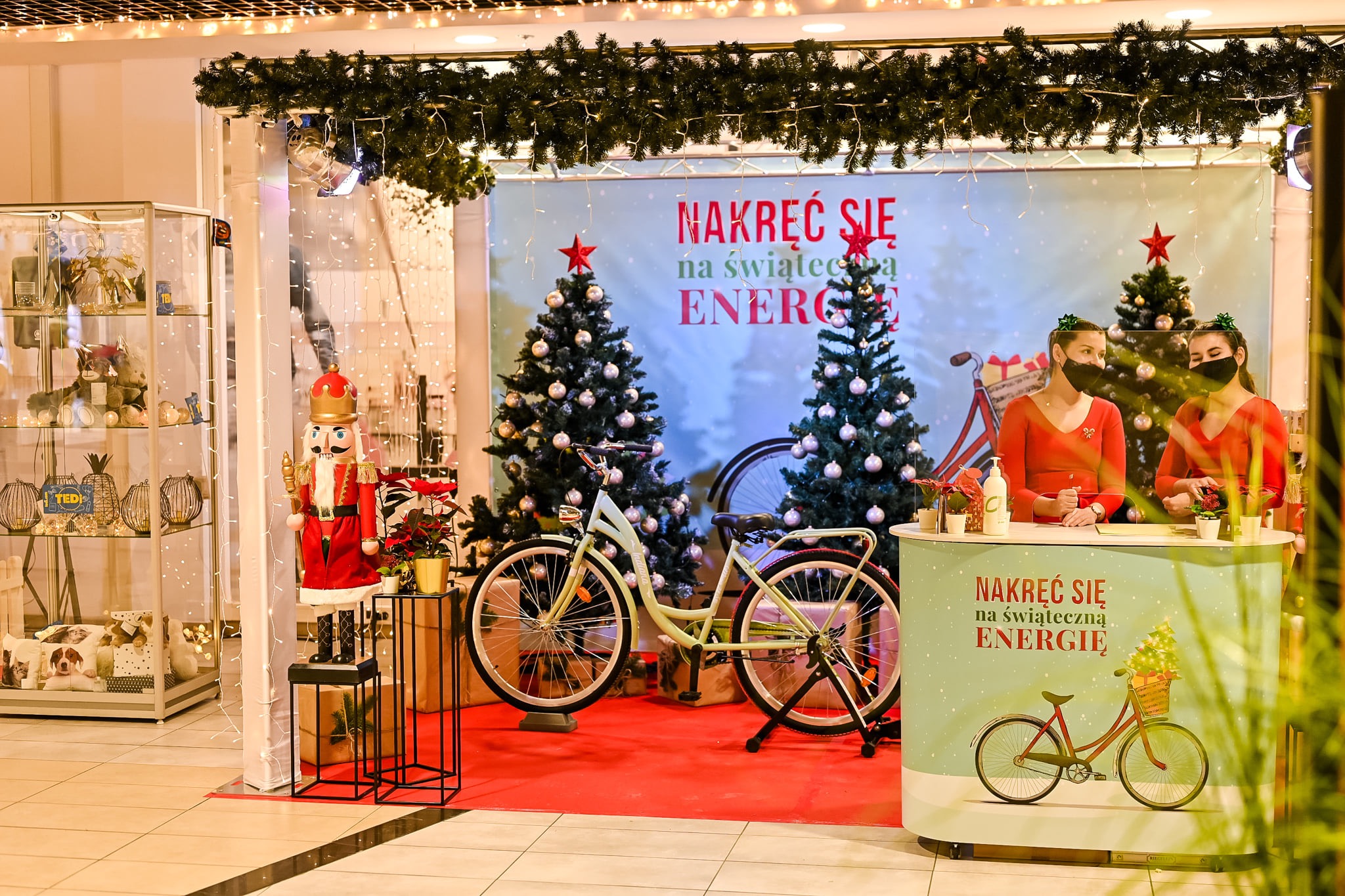 Stoisko promocyjne z dwoma kobietami w czerwonych bluzkach i maseczkach, dekoracjami świątecznymi, rowerem i napisem 'Nakręć się na świąteczną energię'.
