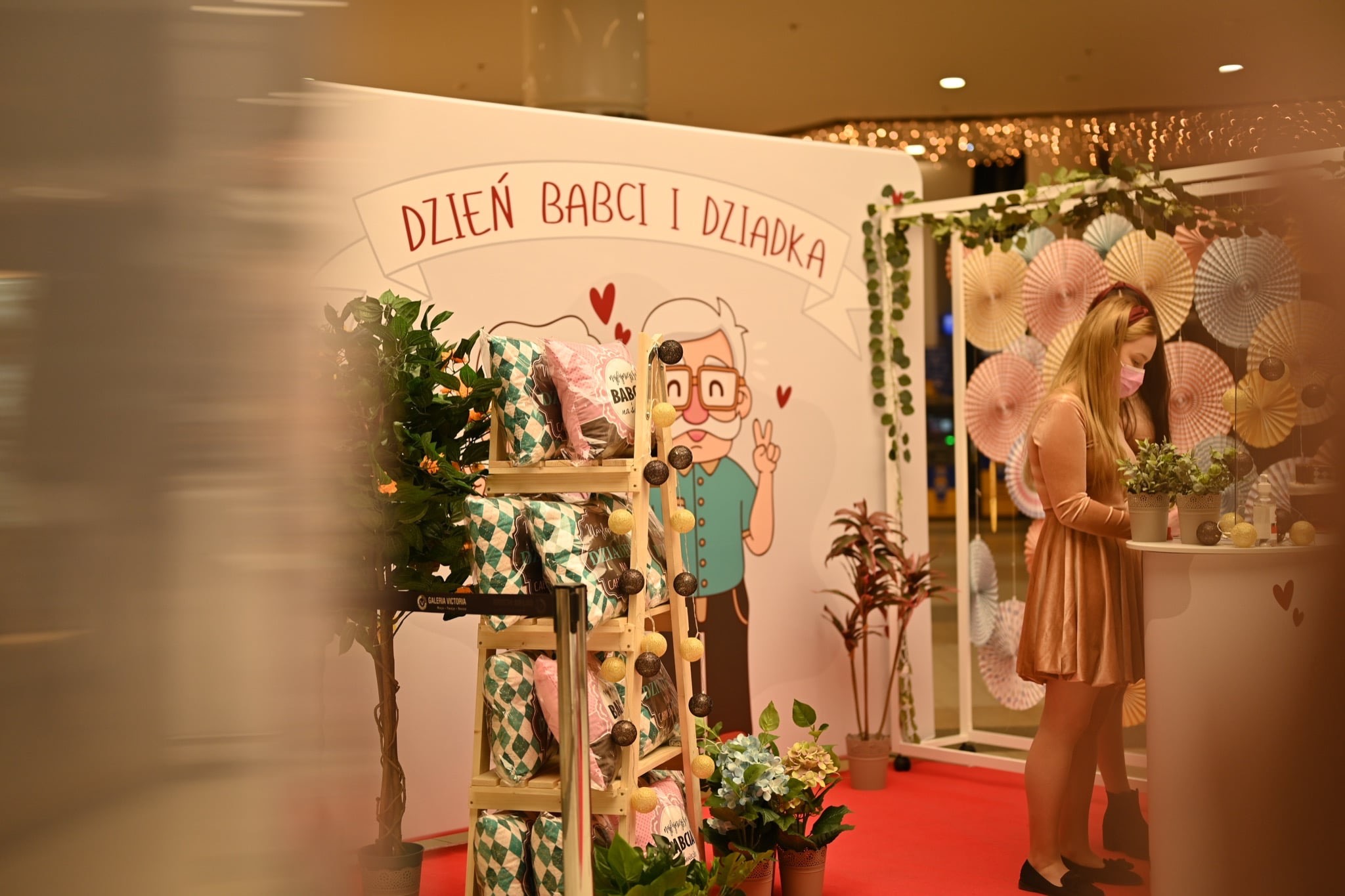 Stoisko z dekoracjami i prezentami z okazji Dnia Babci i Dziadka w centrum handlowym Galeria Victoria, z dziewczyną w maseczce przy ladzie i dekoracyjną ścianką z rysunkiem dziadka.