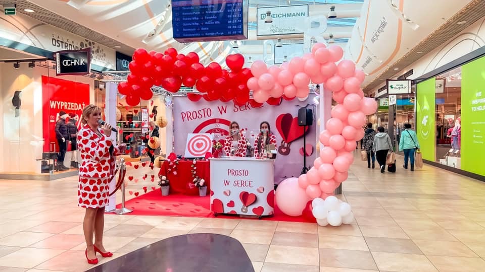 Prezenterka w stroju w serca z mikrofonem podczas wydarzenia promocyjnego 'Prosto w serce' w centrum handlowym, dekoracje z balonów w kształcie serca.