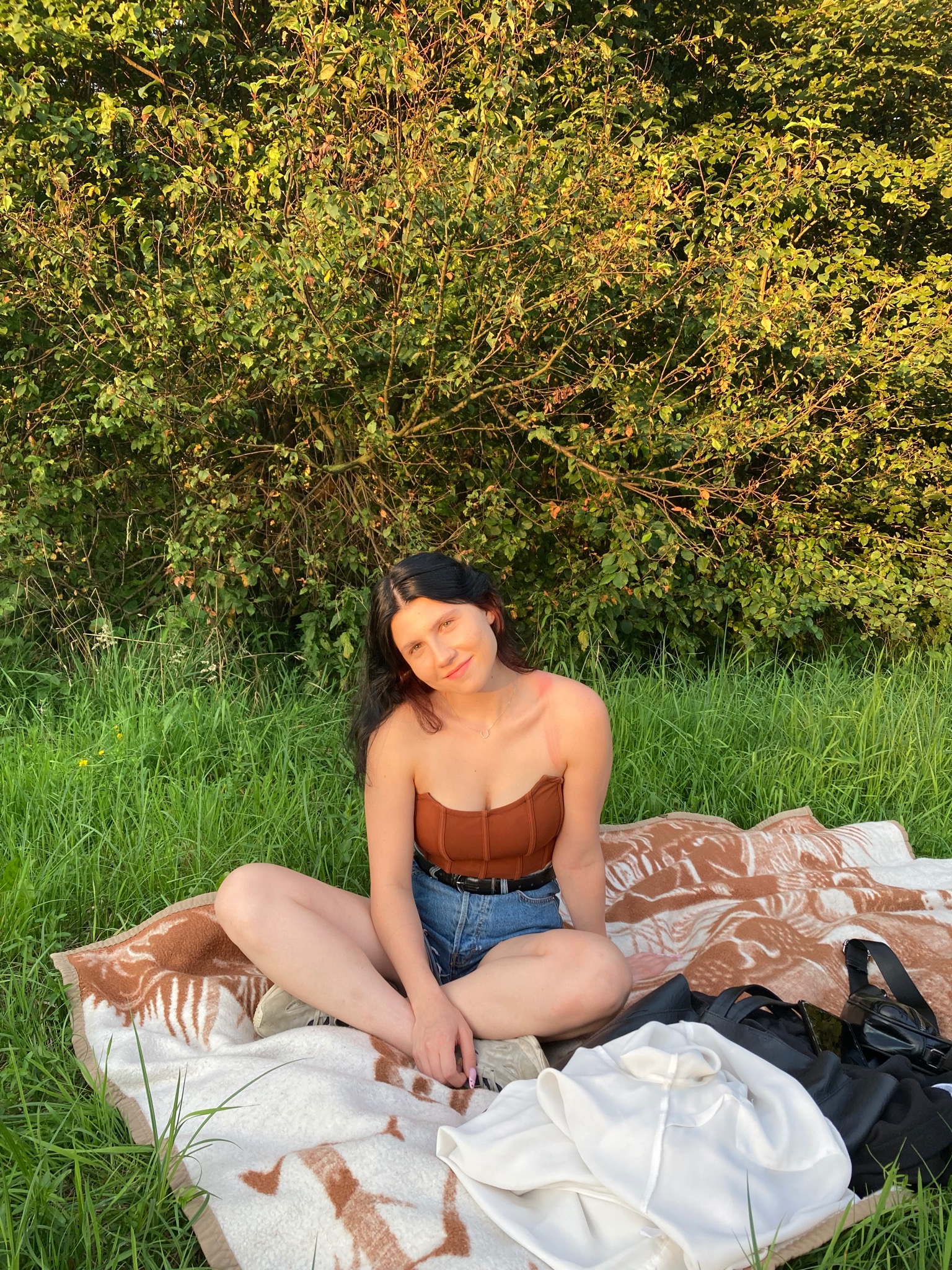 Młoda kobieta w brązowym topie i dżinsowych szortach siedzi na kocu w trawie przed krzakiem, w promieniach zachodzącego słońca.