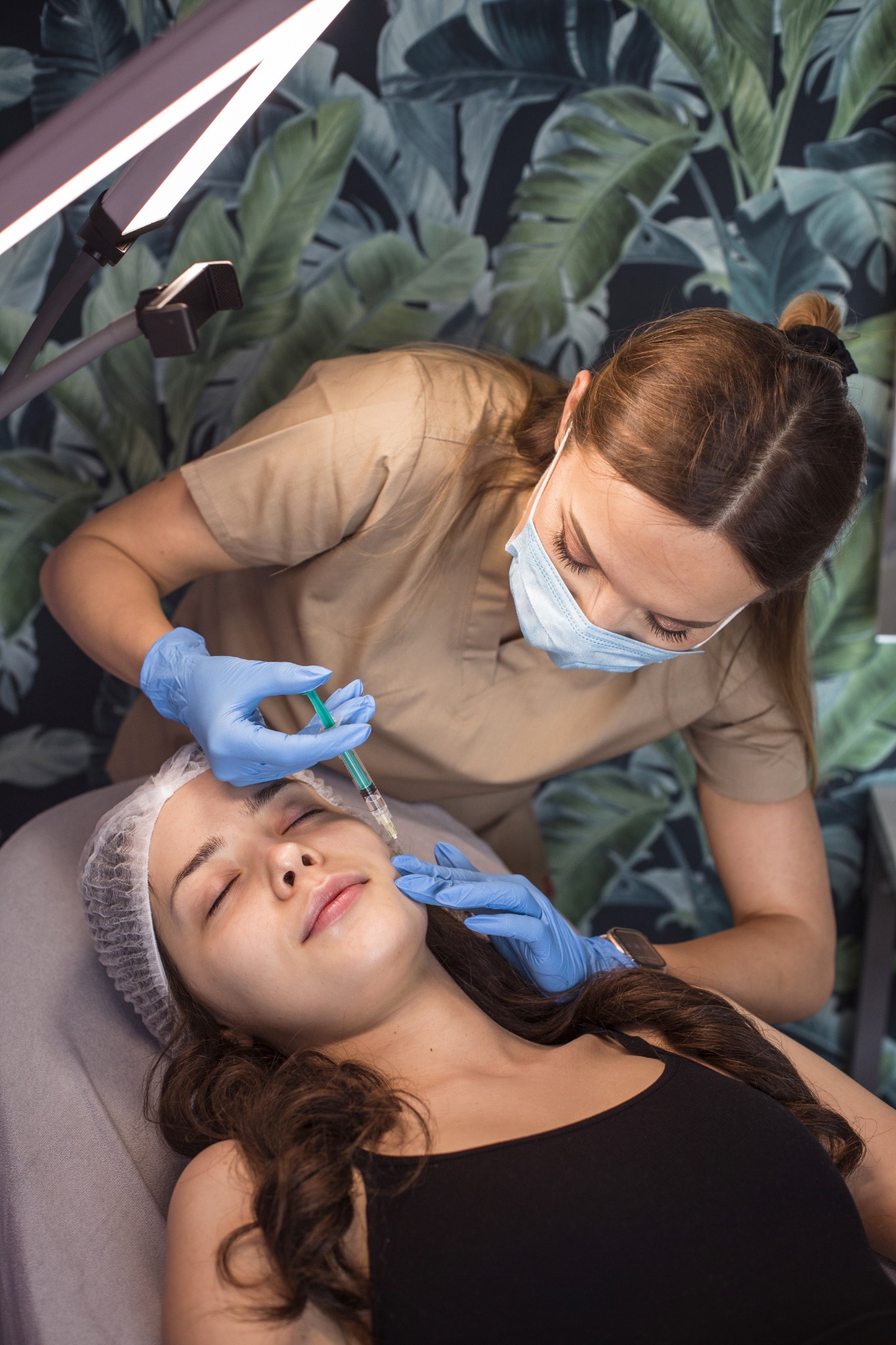 Kobieta w czepku ochronnym leży na leżance podczas zabiegu z użyciem igły, wykonywanego przez kosmetyczkę w rękawiczkach, na tle ściany z motywem roślinnym i pod lampą kosmetyczną.