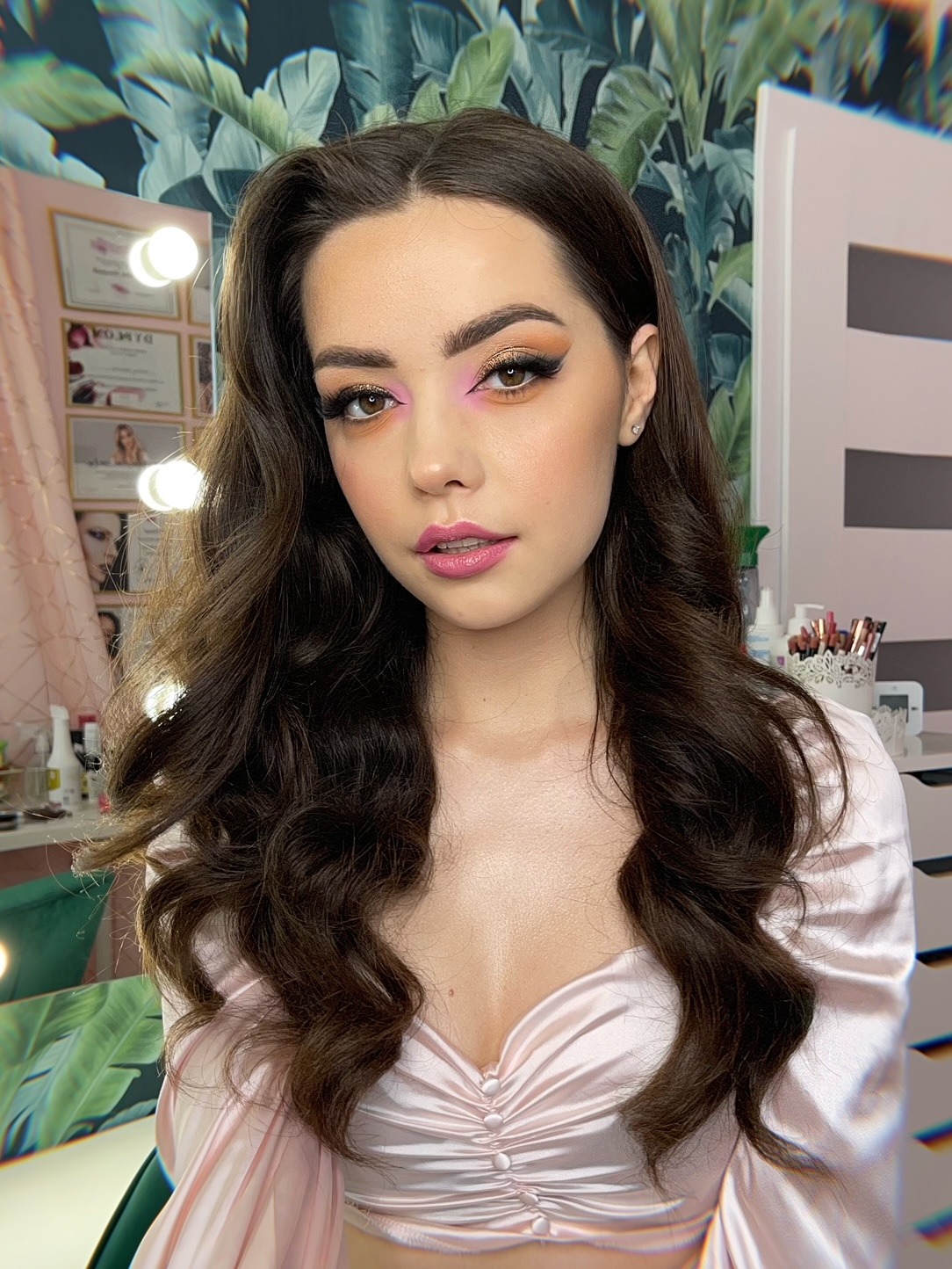 Portret młodej kobiety z profesjonalnym makijażem oczu w odcieniach różu i złota, oraz błyszczącymi ustami w kolorze różowym, z delikatnymi kolczykami w uszach, na tle ściany z motywem liści...