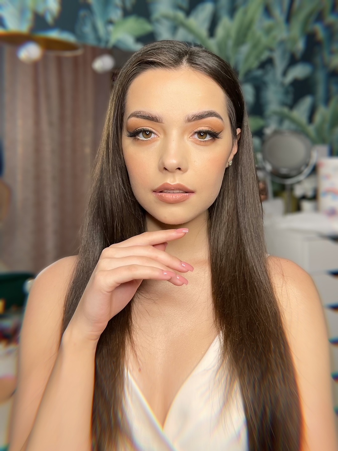 Portret kobiety z delikatnym, brązowym cieniowaniem powiek i podkreślonymi rzęsami, dłoń delikatnie dotyka twarzy, w tle roślinny motyw tapety.