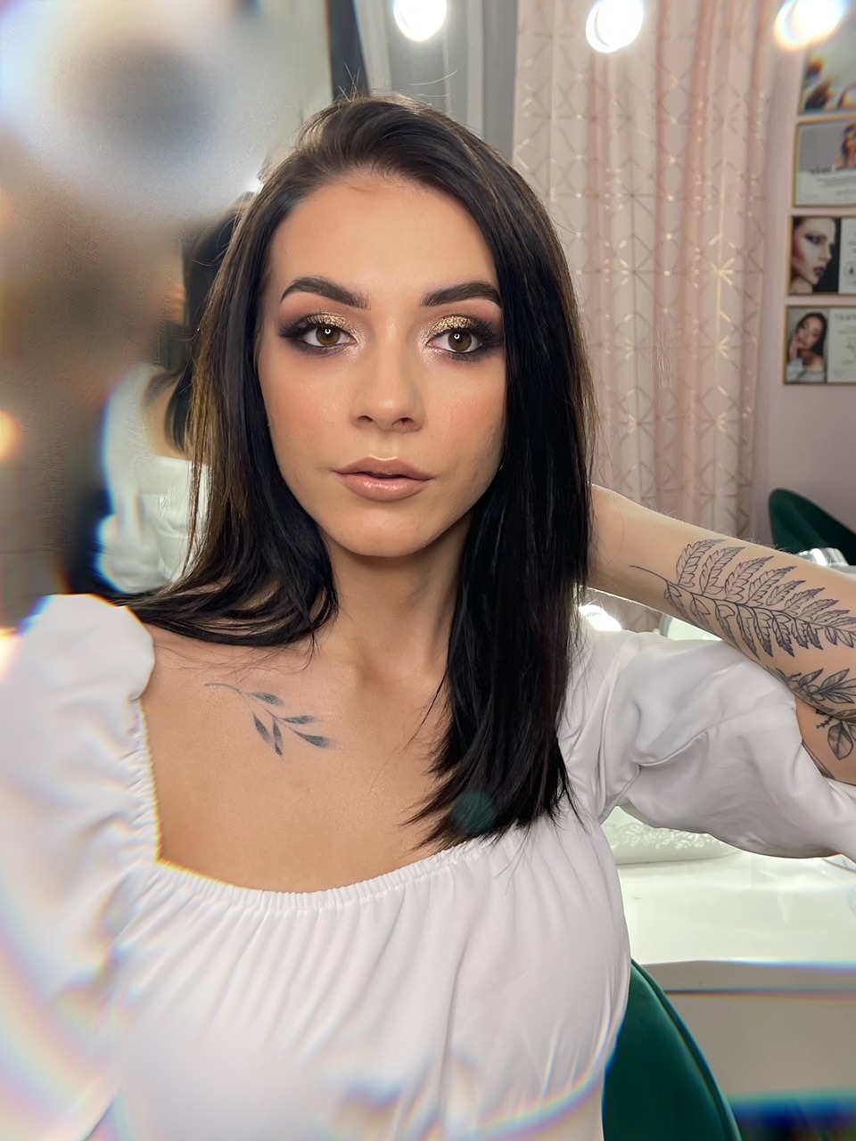 Portret kobiety z ciemnymi włosami i błyszczącym, złotym cieniem na powiekach, wykonanym w Lublinie, z widocznym tatuażem na ramieniu i dekolcie.