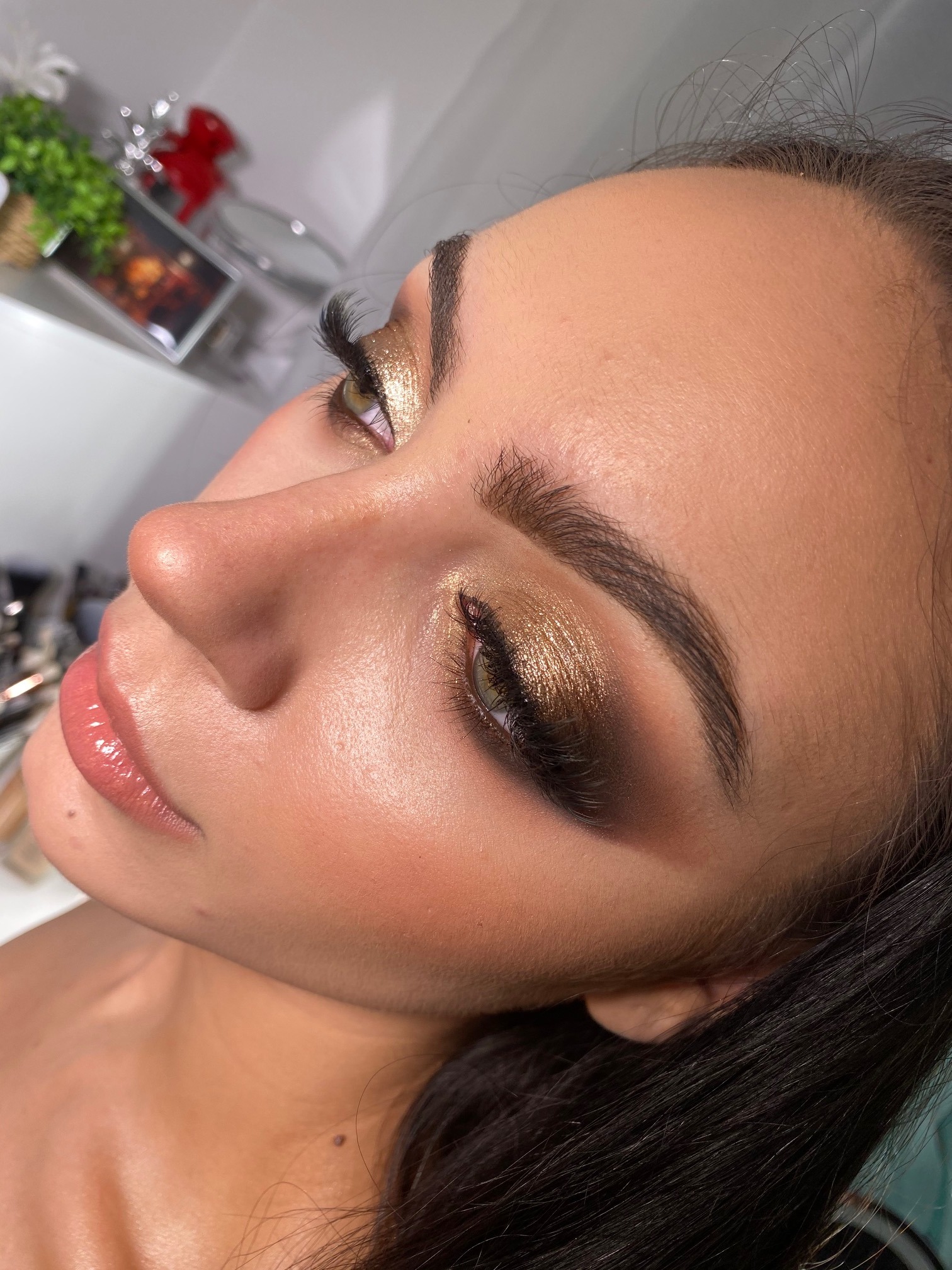 Profil twarzy kobiety z intensywnym, złotym cieniowaniem powiek, długimi sztucznymi rzęsami i błyszczącymi ustami w odcieniu nude, oświetlenie studyjne.